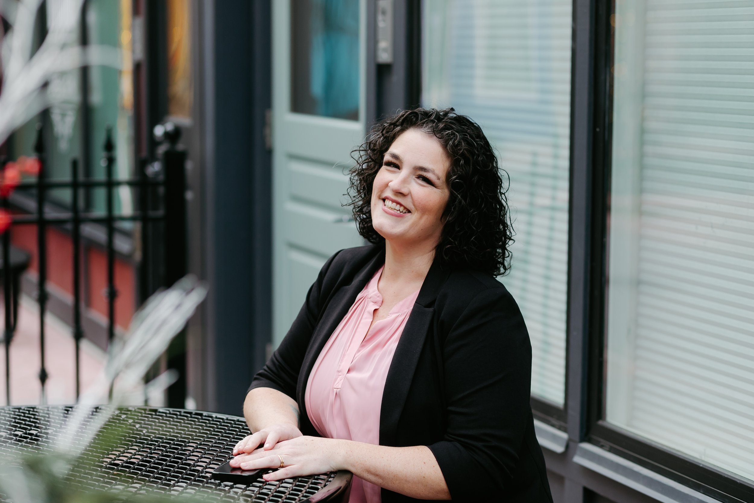 A woman with curly black hair smiling and sitting at an outdoor table.