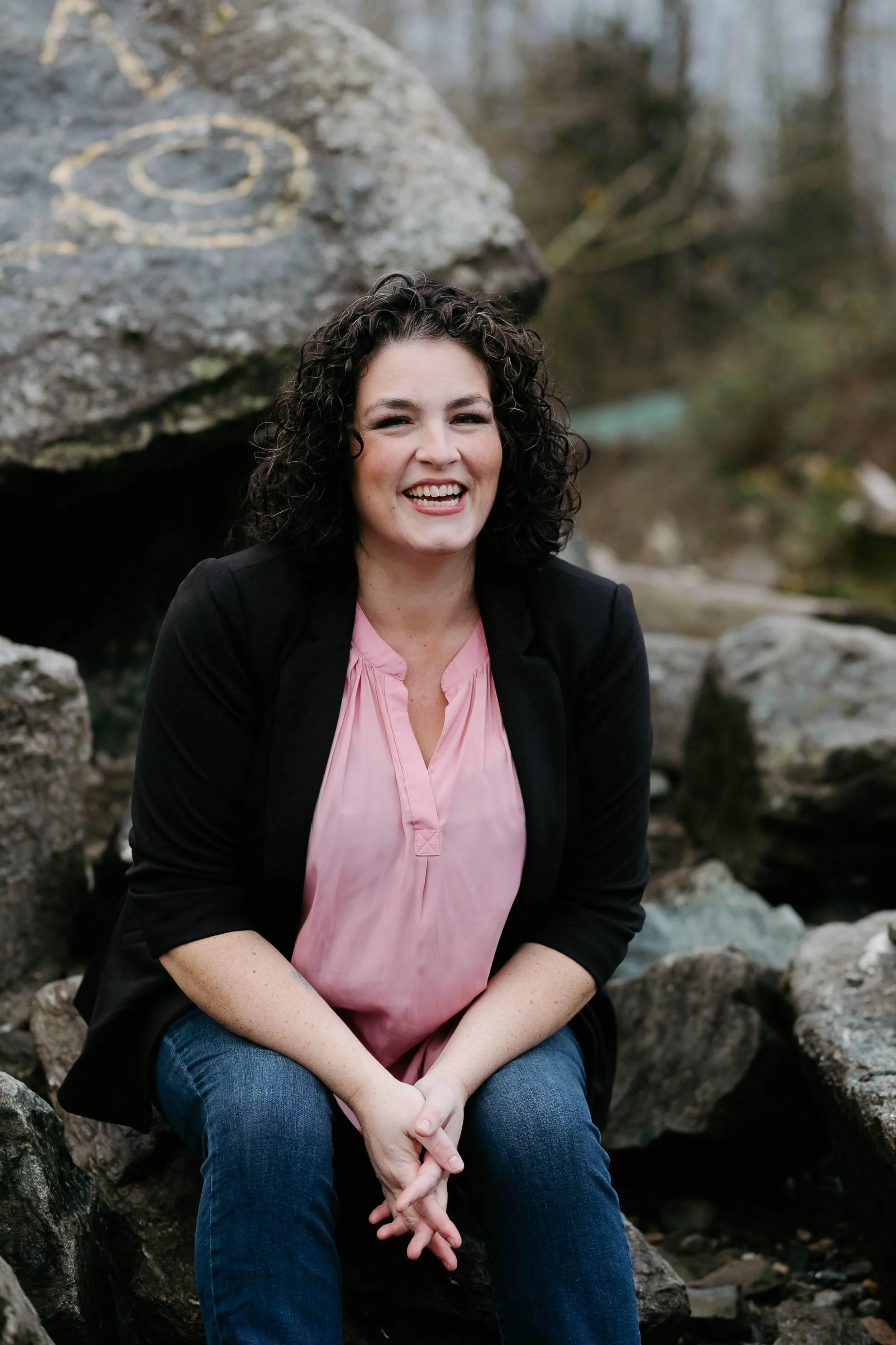 A woman with curly dark hair smiling in an outdoor setting with rocks and water in the background.