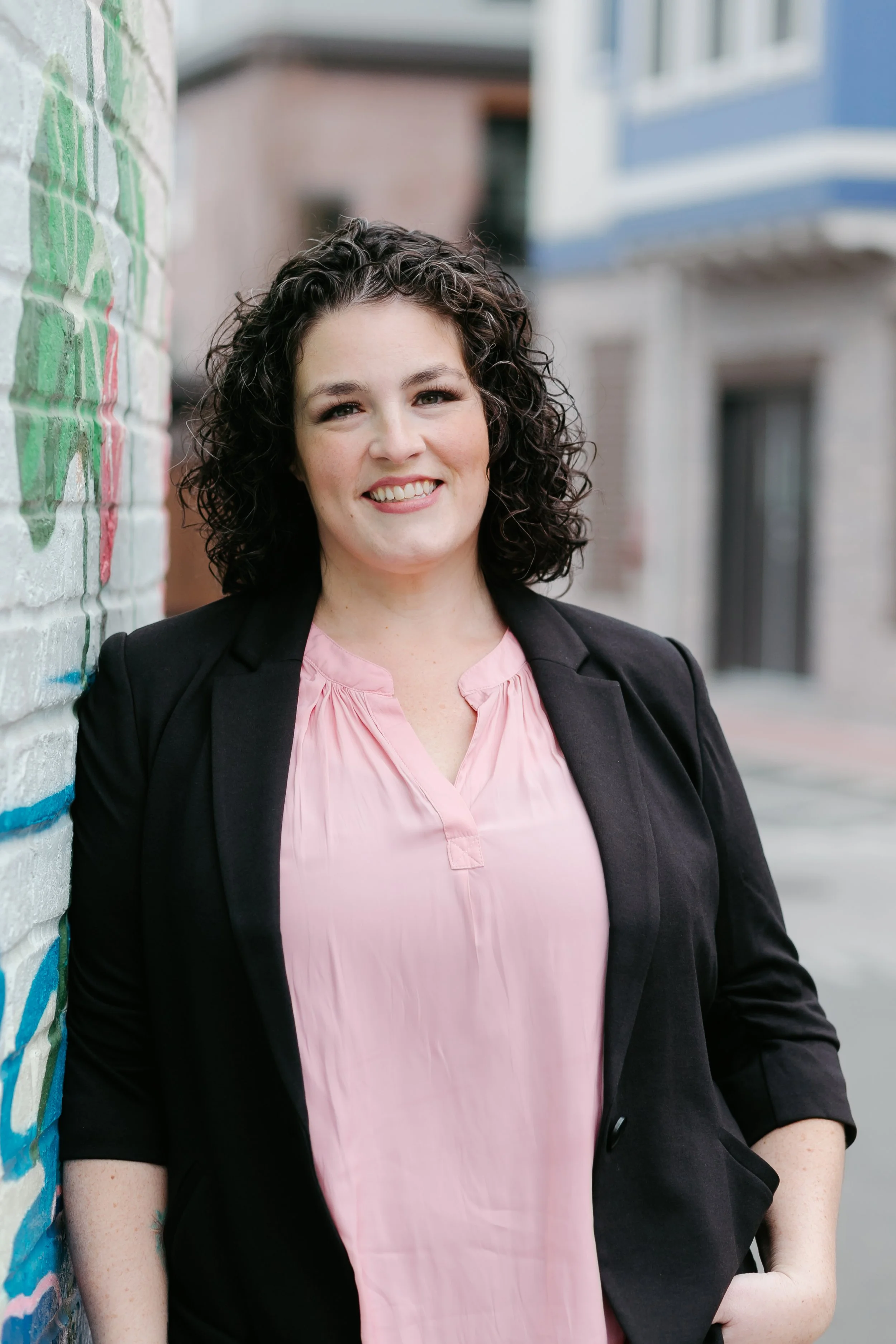 Young woman with curly dark hair smiling, leaning against a graffiti-covered wall in an urban setting.