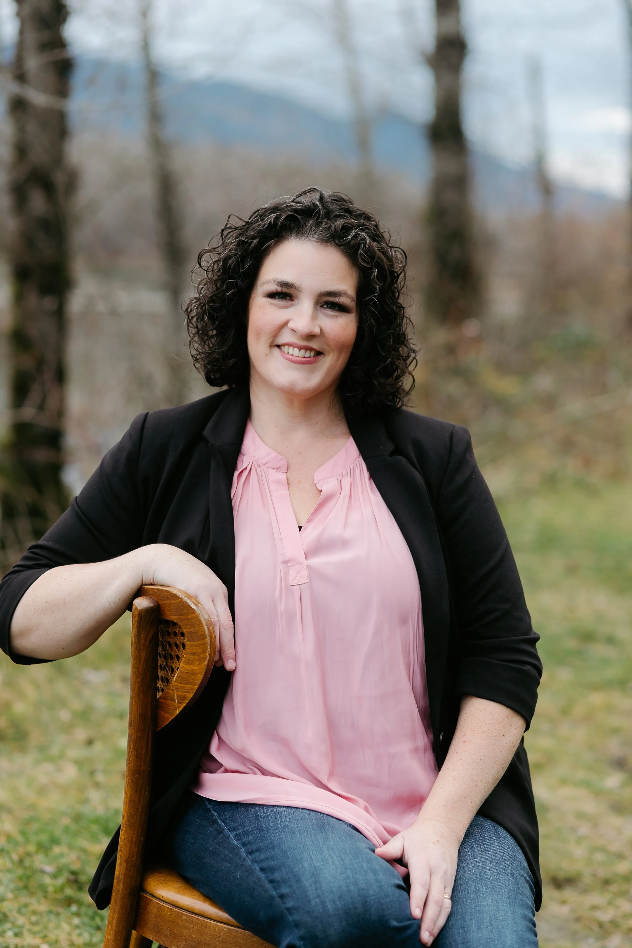 A woman with curly dark hair smiling outdoors, sitting on a wooden chair, wearing a pink blouse and black blazer, in a natural setting with trees and mountains in the background.