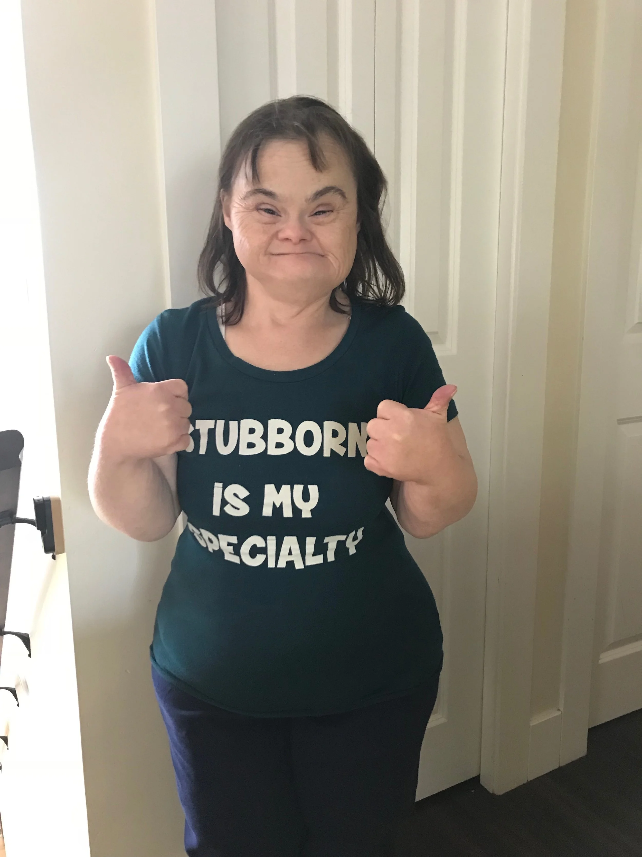 A woman with shoulder-length dark hair and a wide smile, giving two thumbs up, wearing a black T-shirt that says 'STUBBORN IS MY SUPERPOWER' in white letters, standing indoors against a light-colored door and wall.