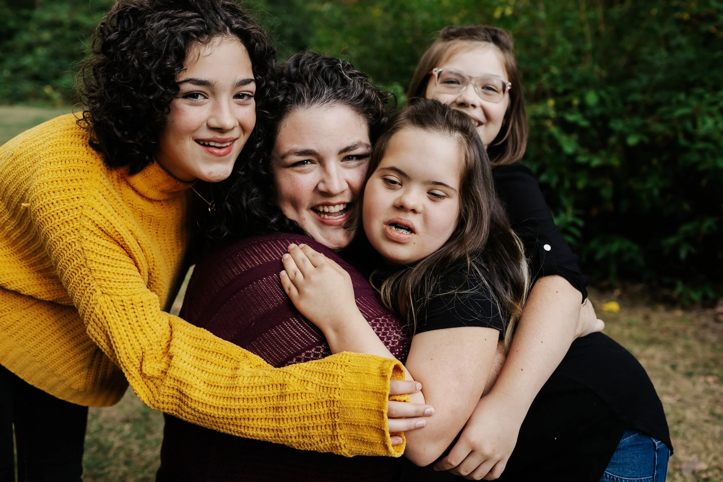 Five young girls hugging outdoors, smiling, and enjoying each other's company, with green foliage in the background.