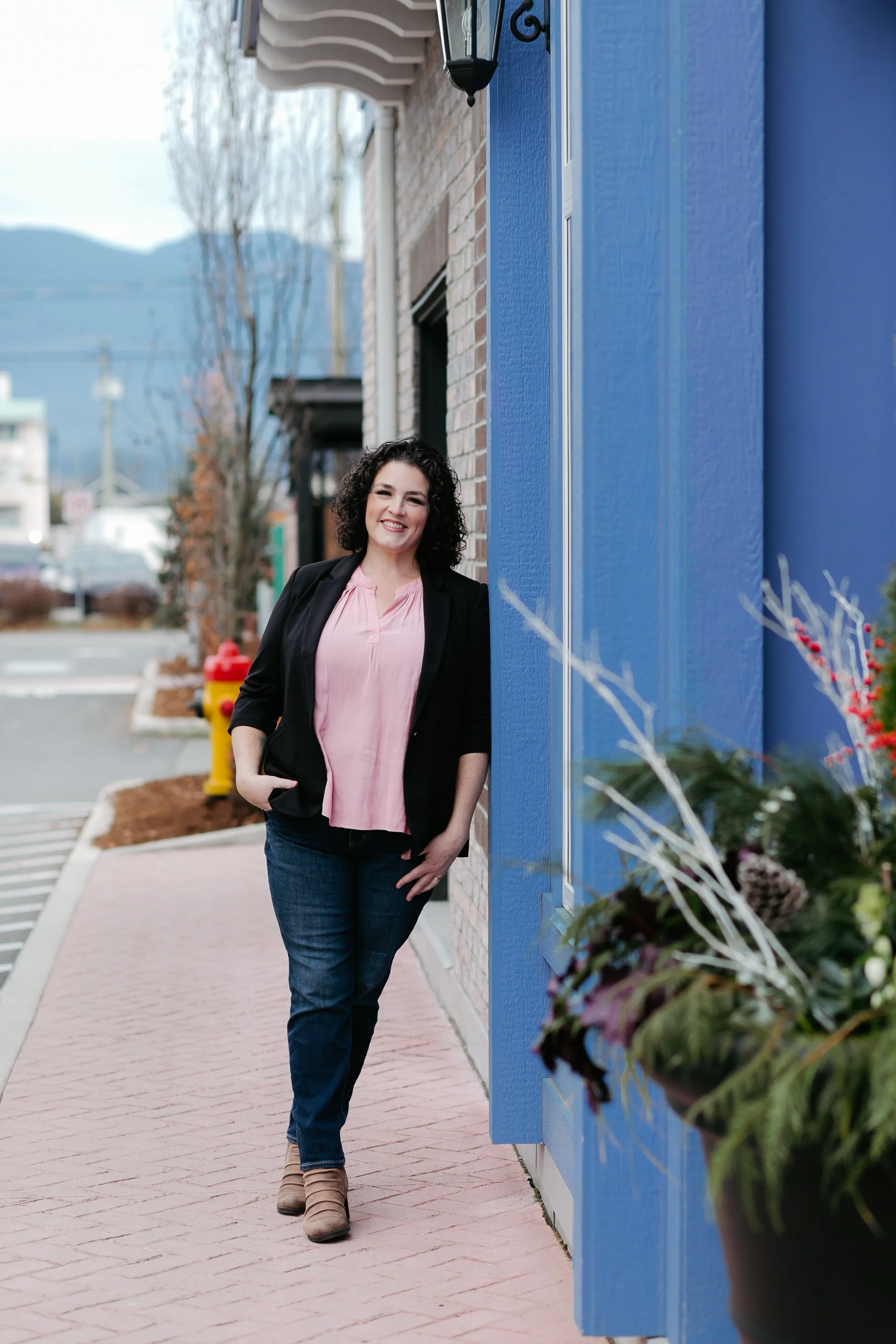 A woman with curly dark hair smiling while standing on a sidewalk next to a blue building with a window and flowerpot.