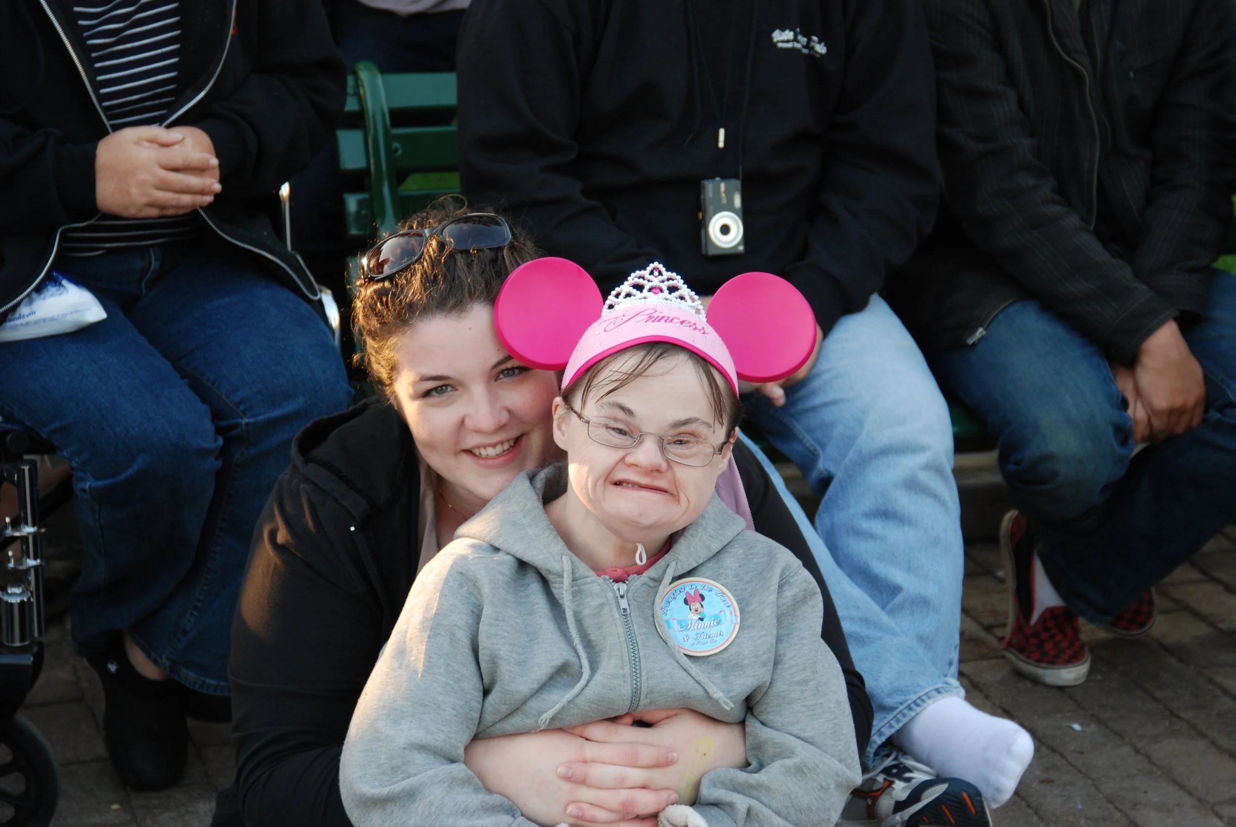 A young woman with sunglasses on her head and a young girl wearing a pink Minnie Mouse ears hat and tiara, smiling together at an outdoor event, surrounded by seated people.
