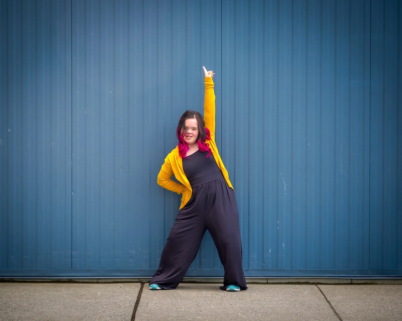 A young woman with pink highlights in her hair, wearing a yellow jacket over a black jumpsuit, striking a confident pose with one arm raised and one hand on her hip, standing in front of a blue metal wall.