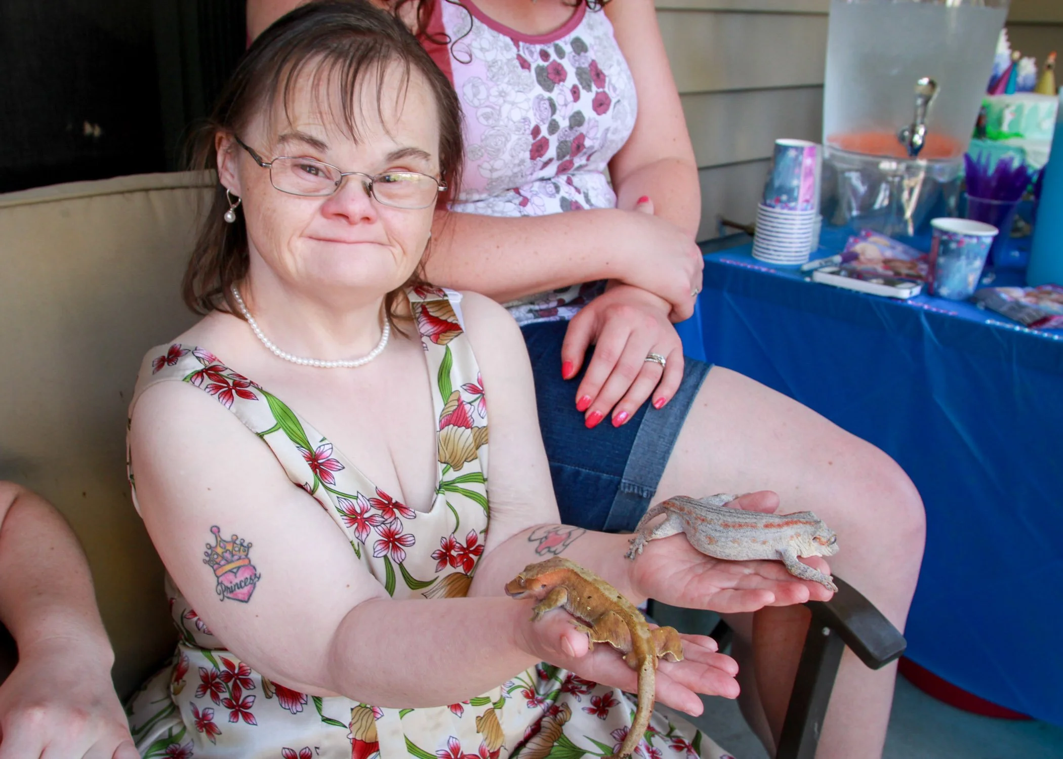 A woman holding two geckos, one on each hand, at a celebration with a blue tablecloth and party supplies in the background.