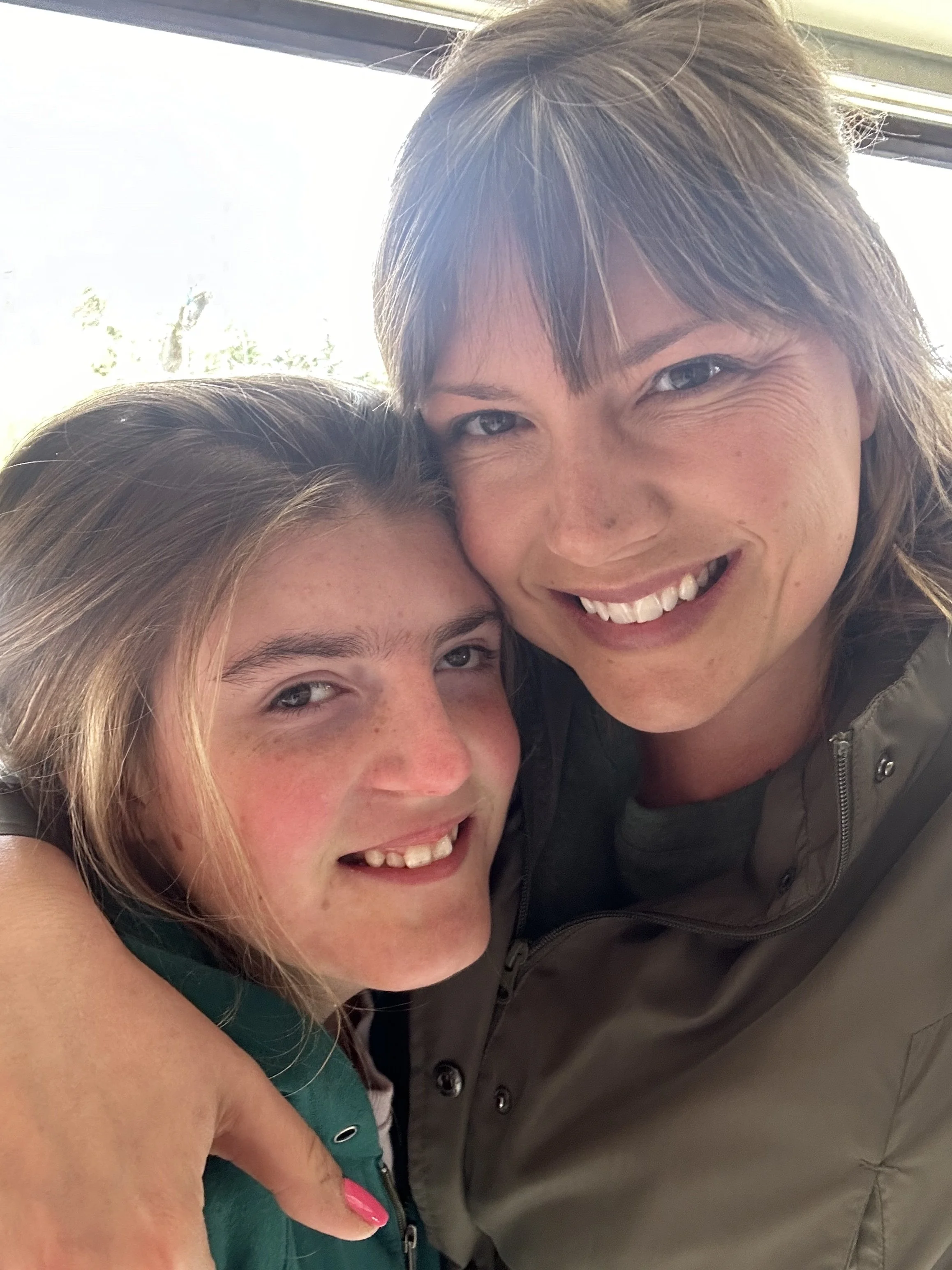 A woman and a girl are hugging and smiling at the camera with a bright background.