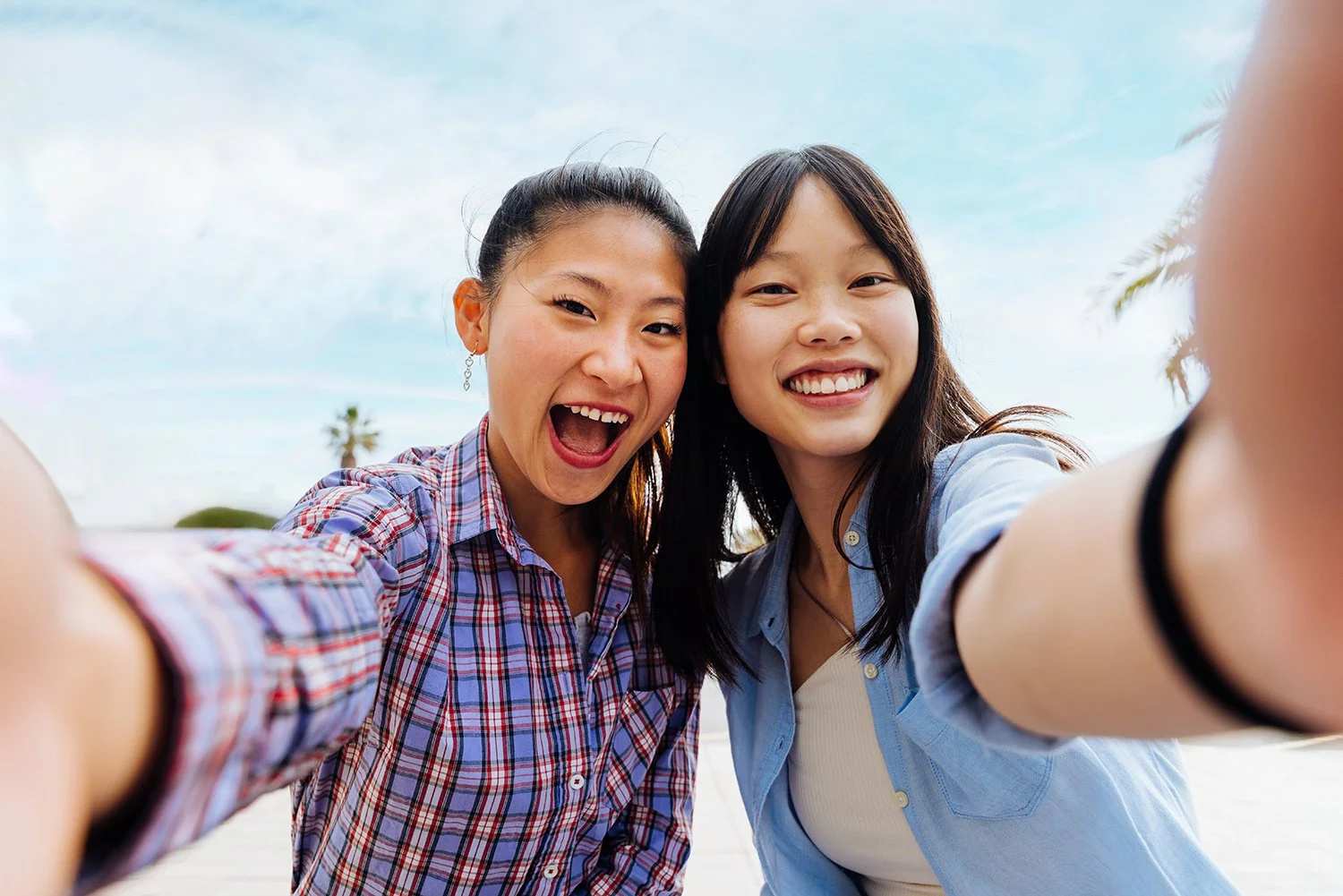 Two smiling young women taking a selfie outdoors with a bright sky and palm trees in the background.
