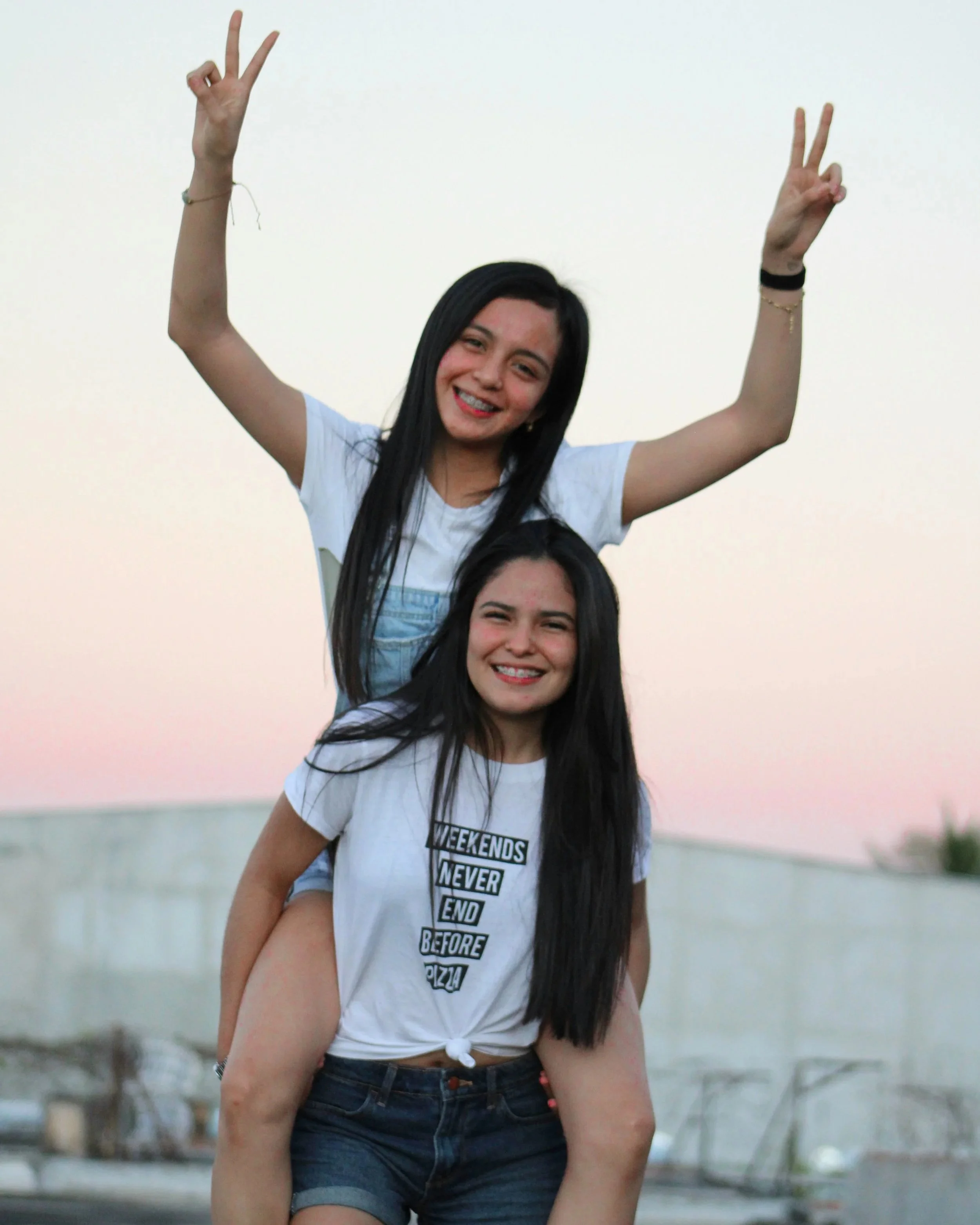 Two young women smiling, one giving a peace sign with both hands, standing outdoors at sunset.