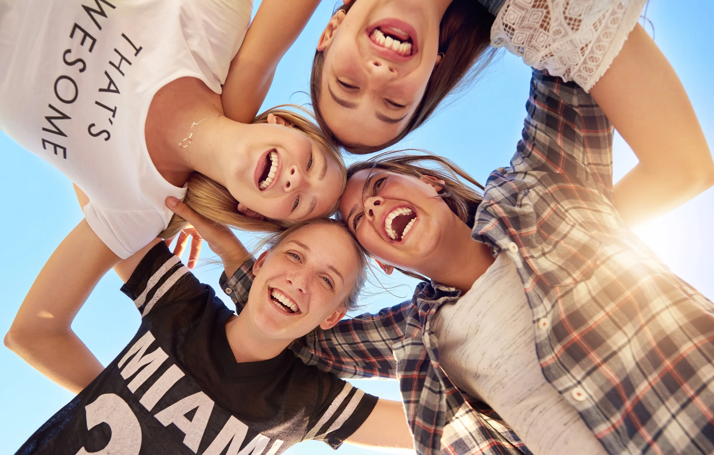 Group of five young women, smiling and laughing, forming a circle with their heads touching, taken from below against a clear blue sky.