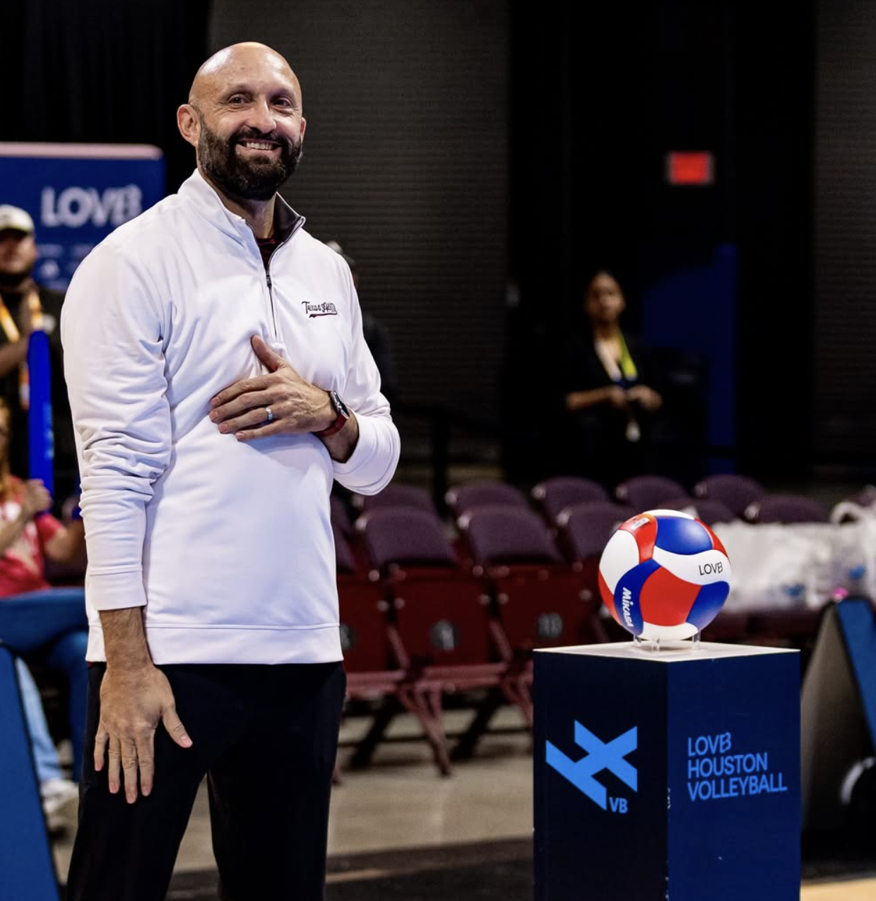 Texas A&M head coach Jamie Morrison wearing a white Texas Strong jacket standing next to a volleyball on a stand with Houston Volleyball and LOVB logos.