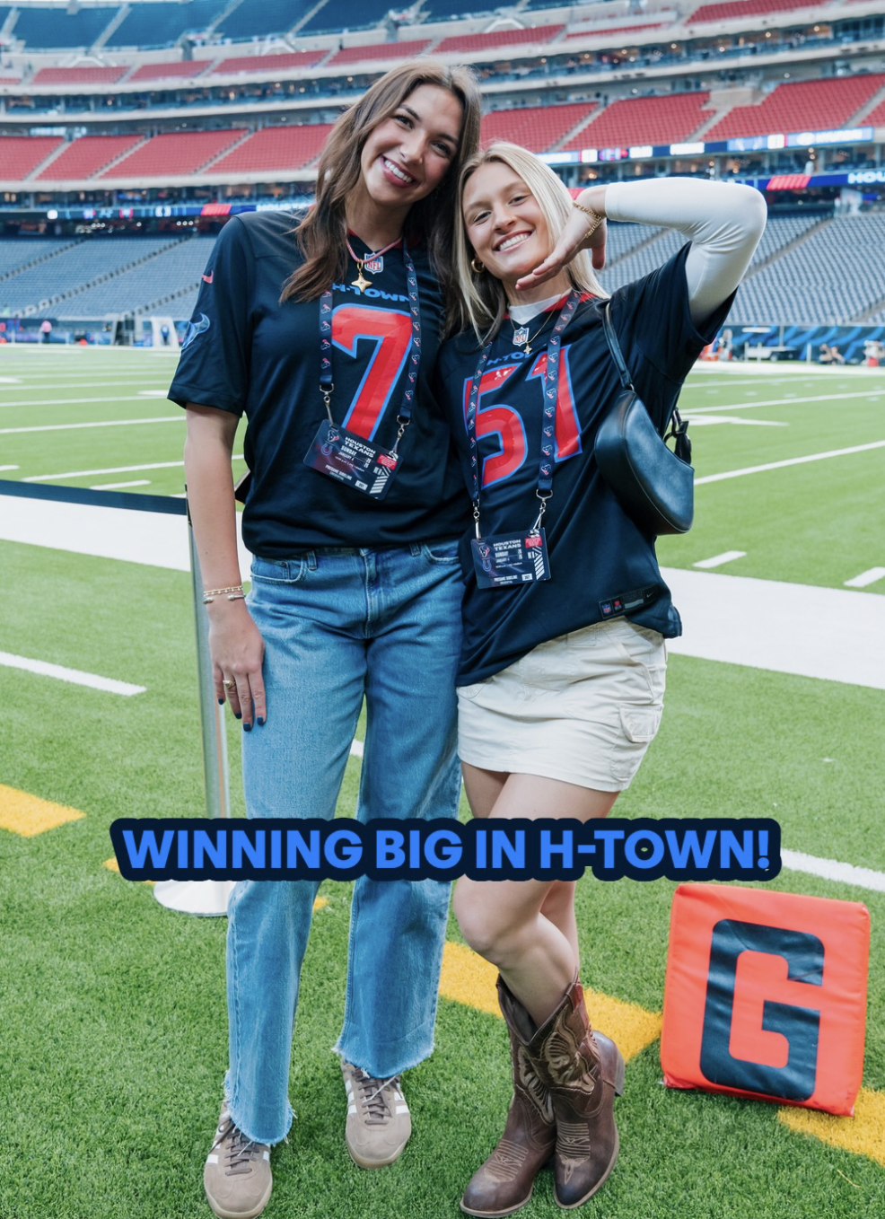 Two smiling women in football jerseys standing on a stadium field, celebrating a game. One woman has short blonde hair, wearing a black shirt with the number 51. The other woman has long brown hair, wearing a black shirt with the number 7. They are wearing event passes around their necks. There is a football field and stadium seating in the background, and a red bag with a G on it on the ground. The text "WINNING BIG IN H-TOWN!" is overlaid at the bottom of the image.