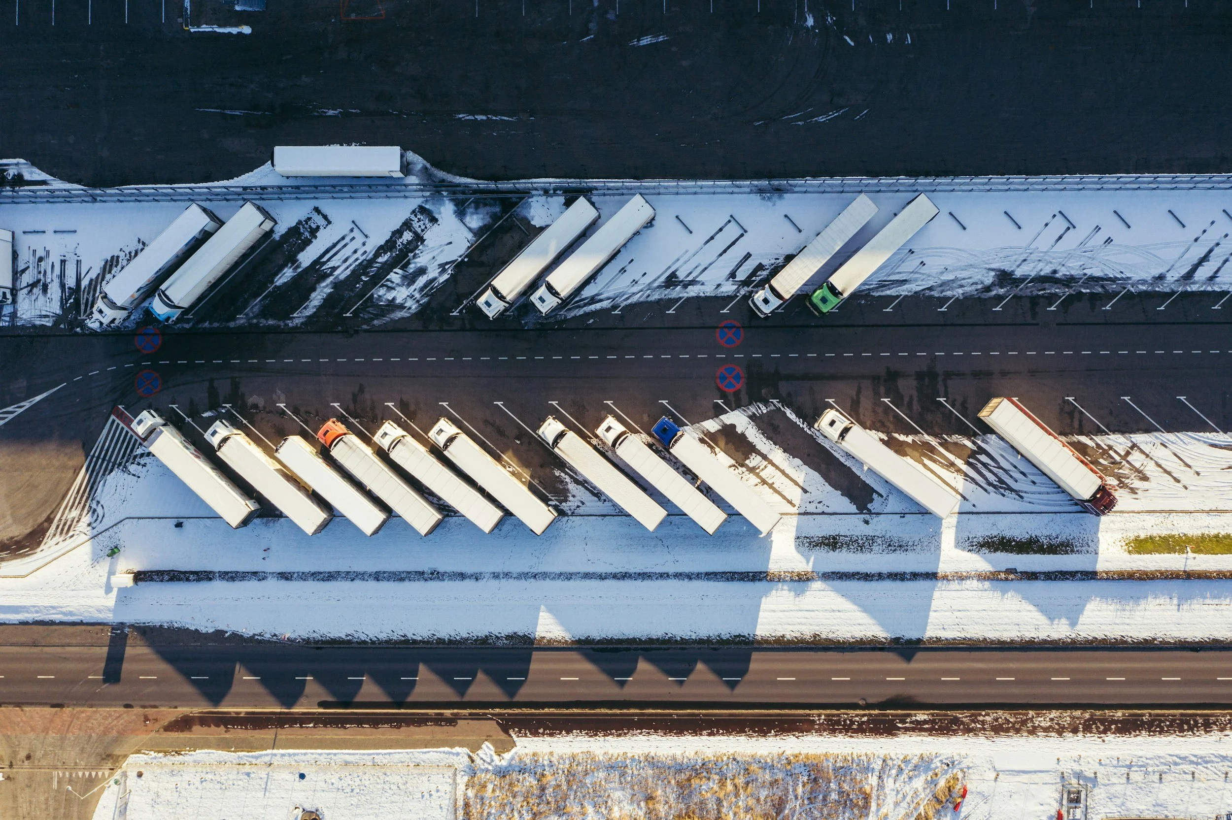 Aerial view of a snow-covered parking lot with trucks parked along the side, next to a building, and a road running parallel at the bottom.