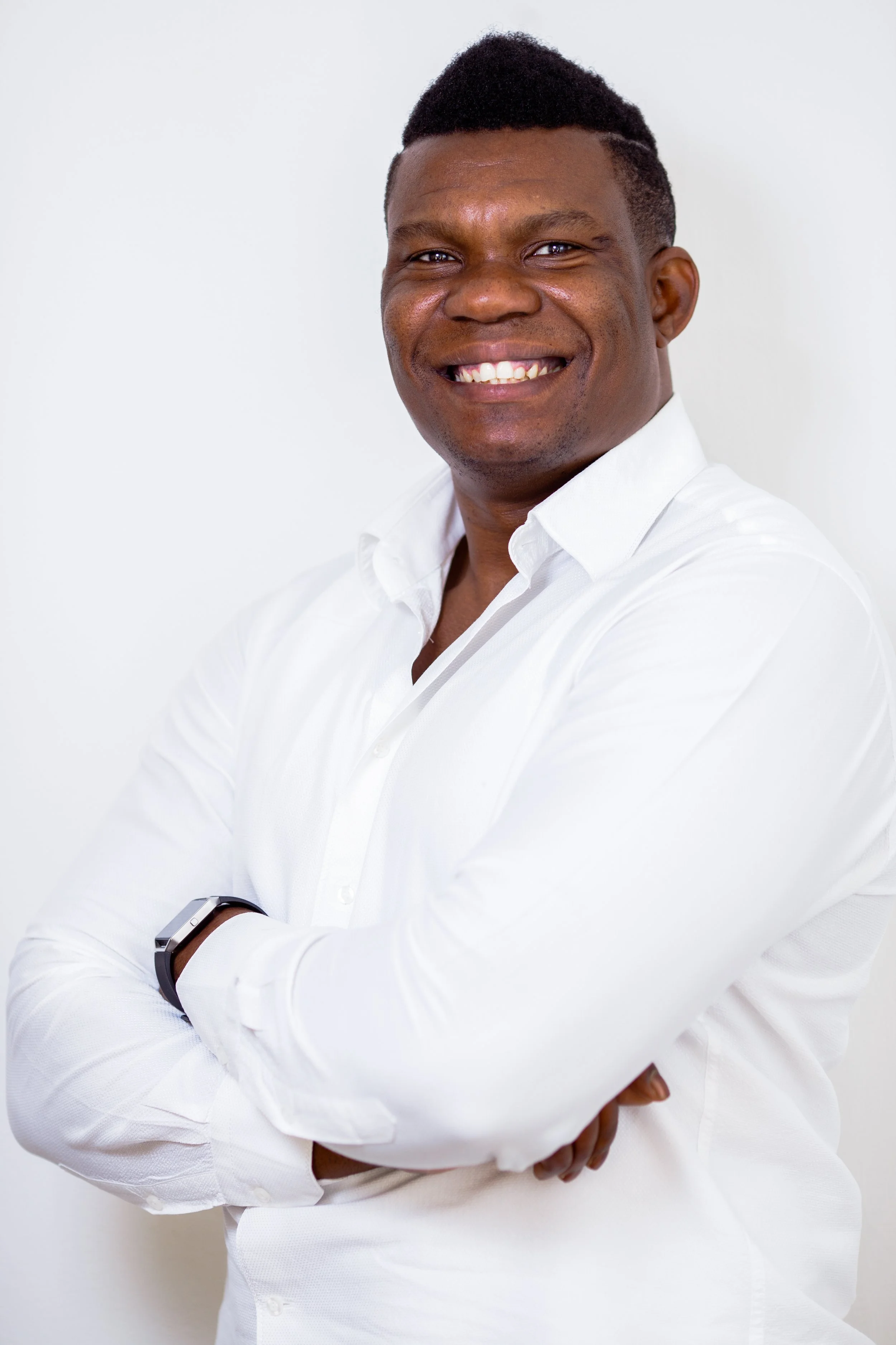 A smiling man with crossed arms wearing a white dress shirt and a wristwatch, standing against a plain white background.