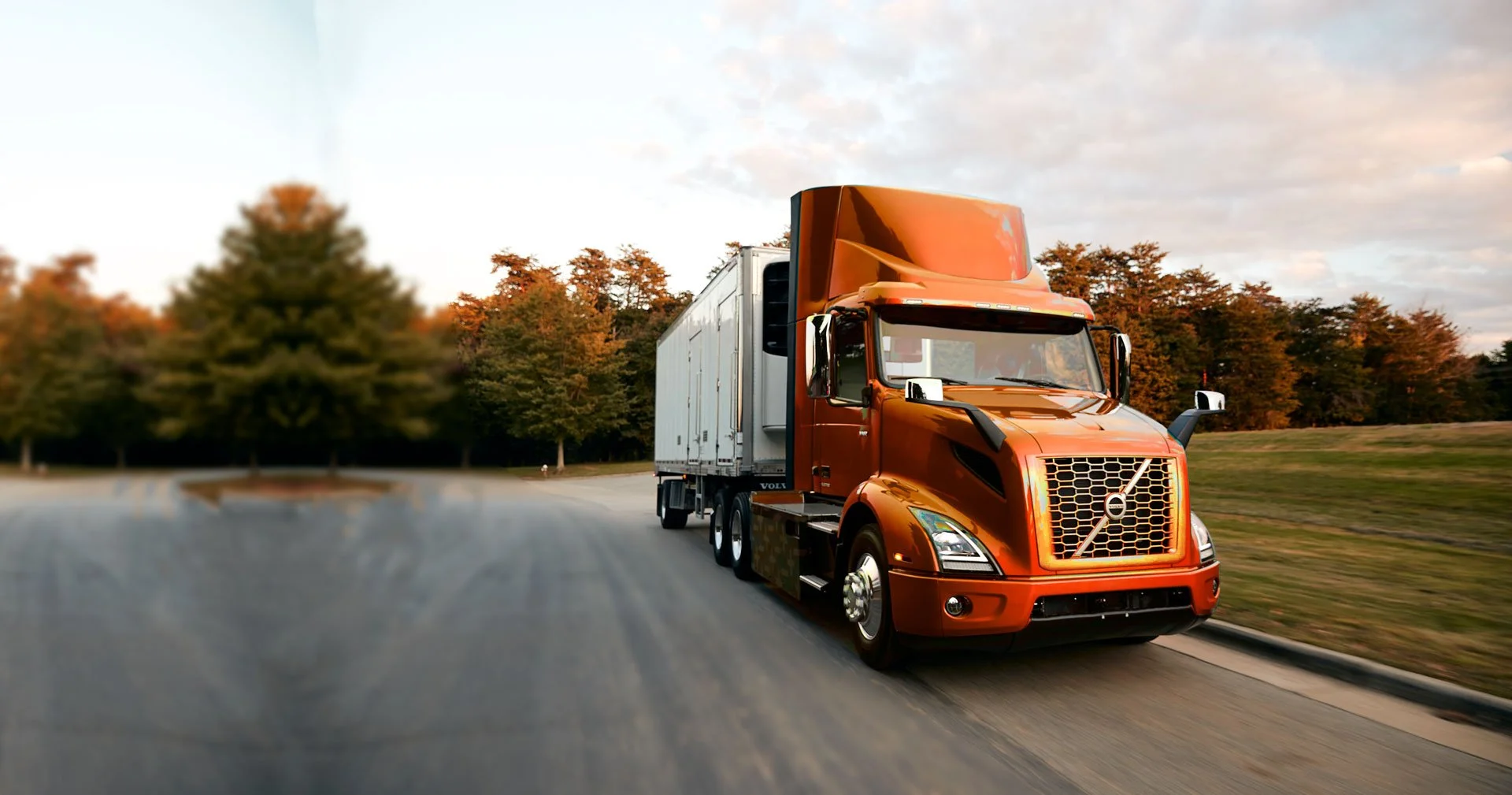 Orange semi-truck driving on a road with trees and a partly cloudy sky in the background.
