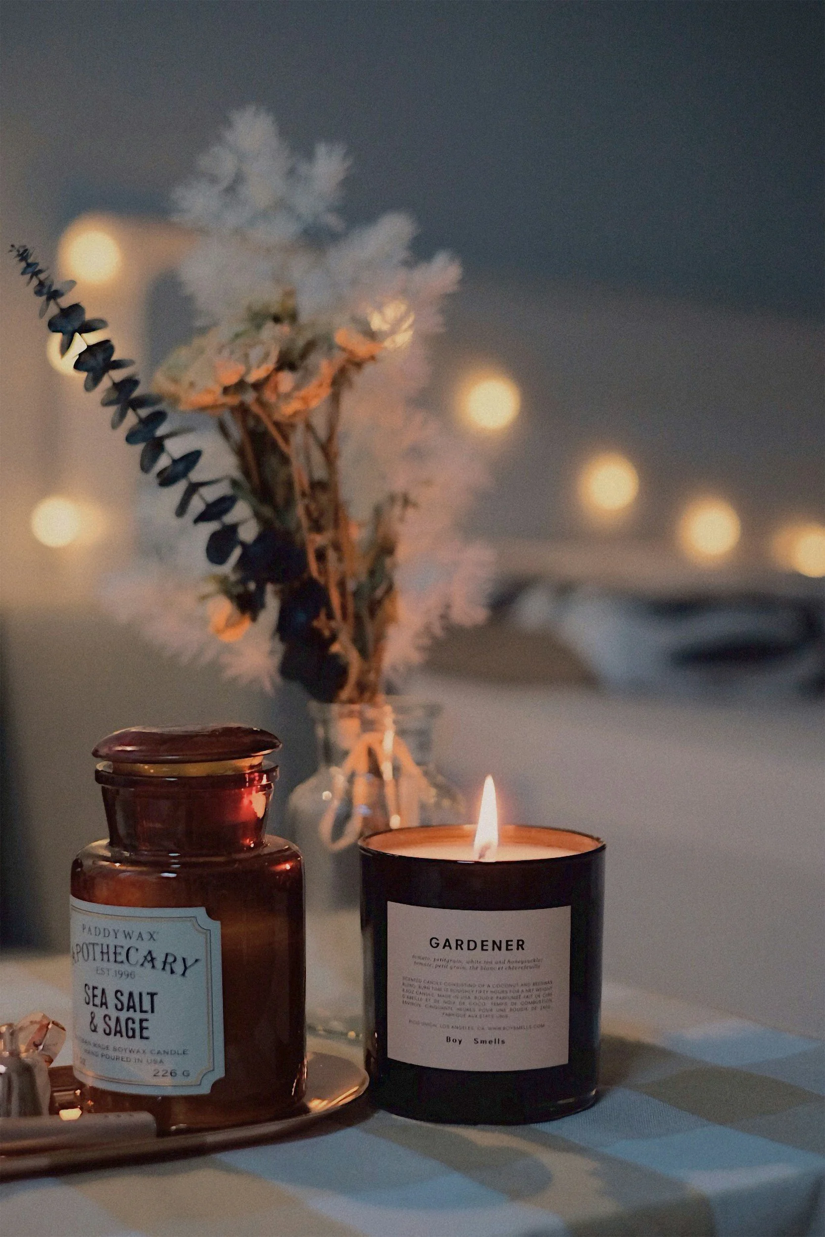 A lit candle labeled 'GARDENER' next to a brown jar labeled 'SEA SALT & SAGE' on a tray, with a vase of dried flowers and eucalyptus in the background, and blurred warm lights in the background.
