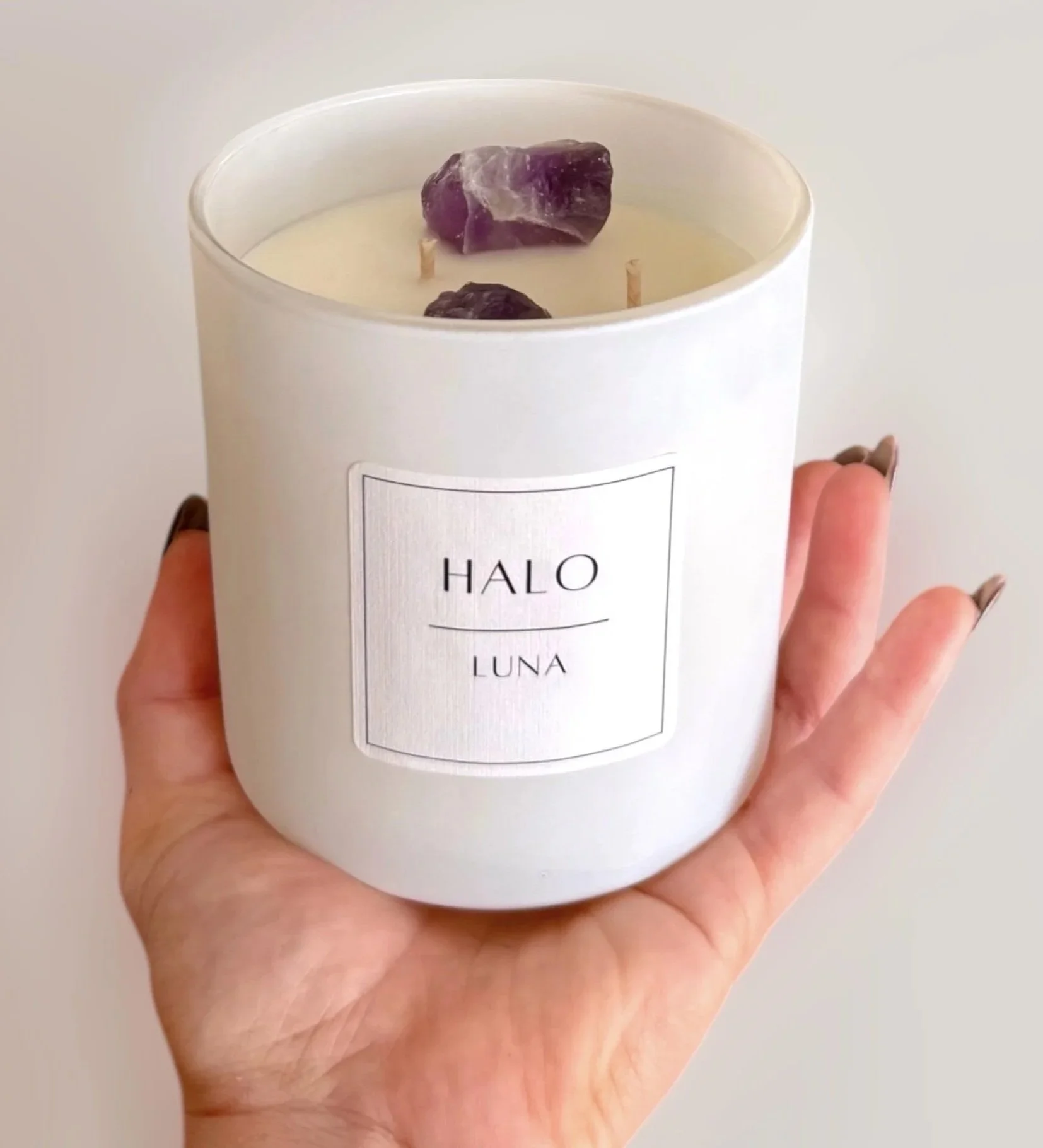 A white candle with purple amethyst crystals inside, held in a person's hand. The candle has a label that reads 'HALO LUNA'.