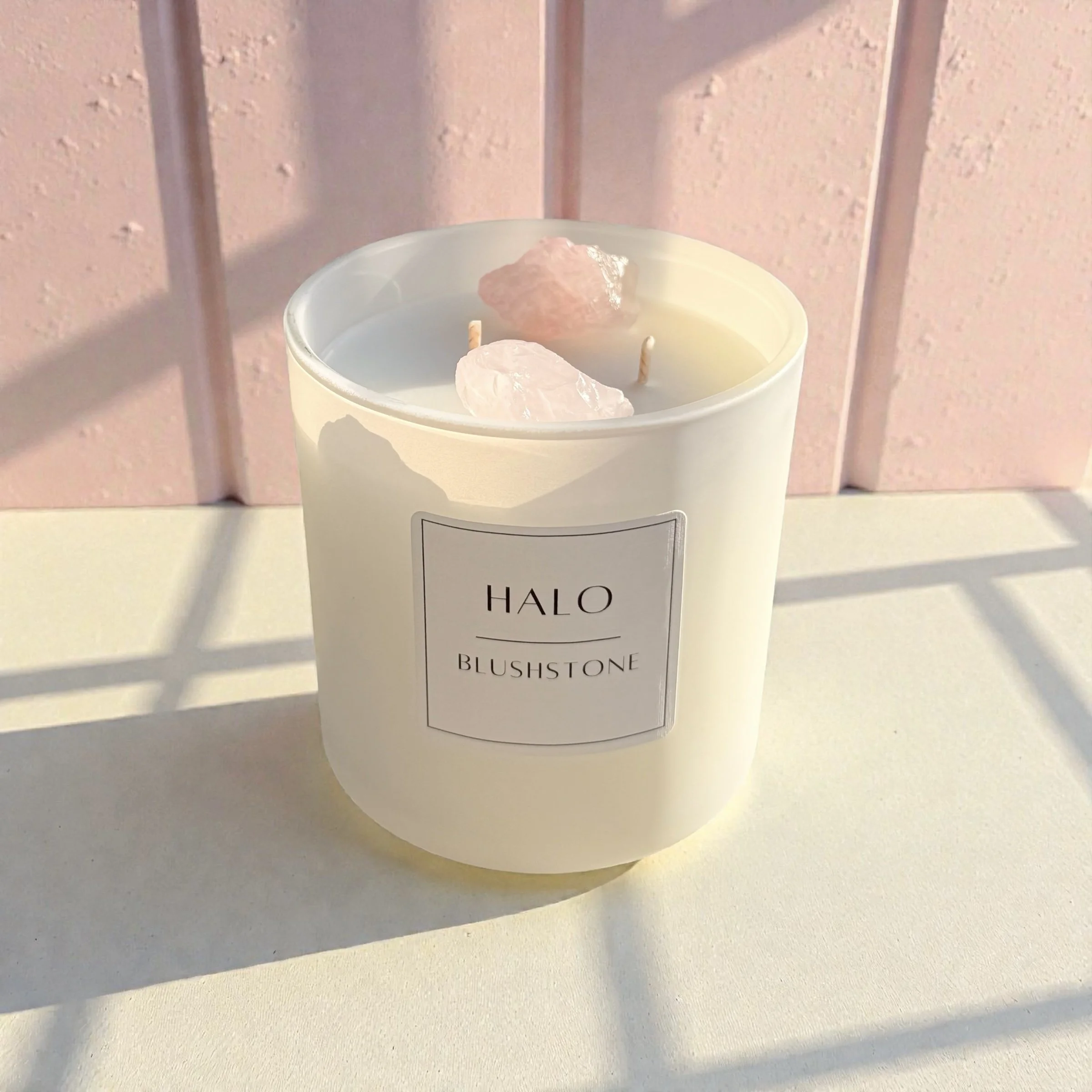 Cream-colored candle labeled 'HALO BLUSHSTONE' with pinkish-white quartz crystals inside, placed on a white surface with sunlight and shadow patterns, pink wall in background.