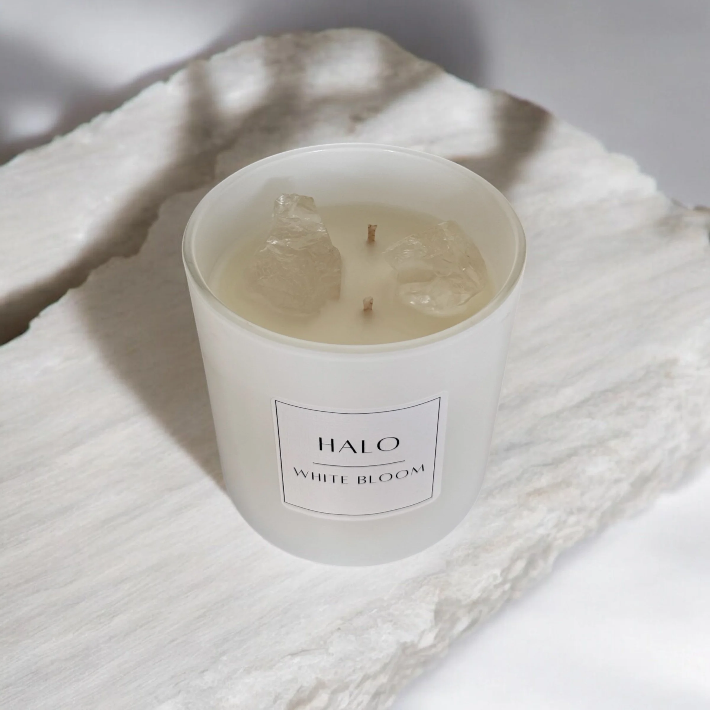 A white scented candle labeled 'HALO White Bloom' with three large clear quartz crystals inside, placed on a textured white stone surface.