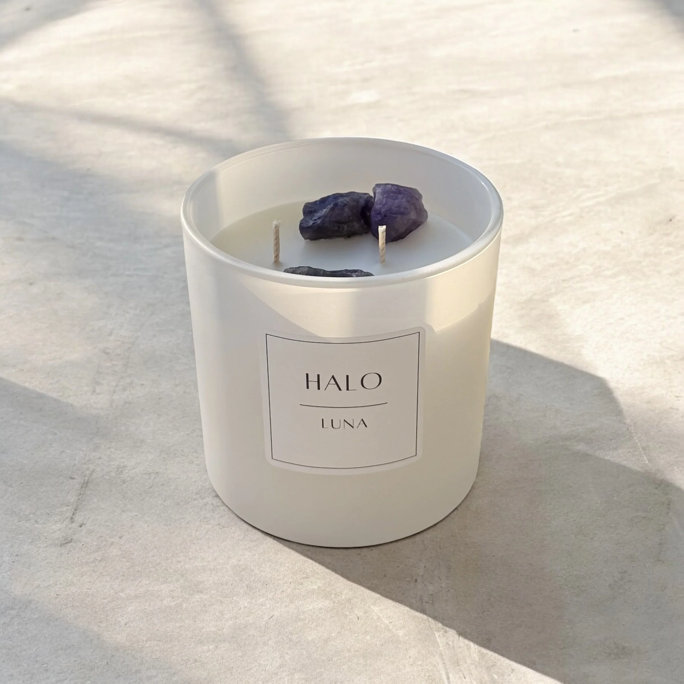 White candle in a glass container labeled 'HALO LUNA' with three purple amethyst crystals on top, placed on a light-colored surface.