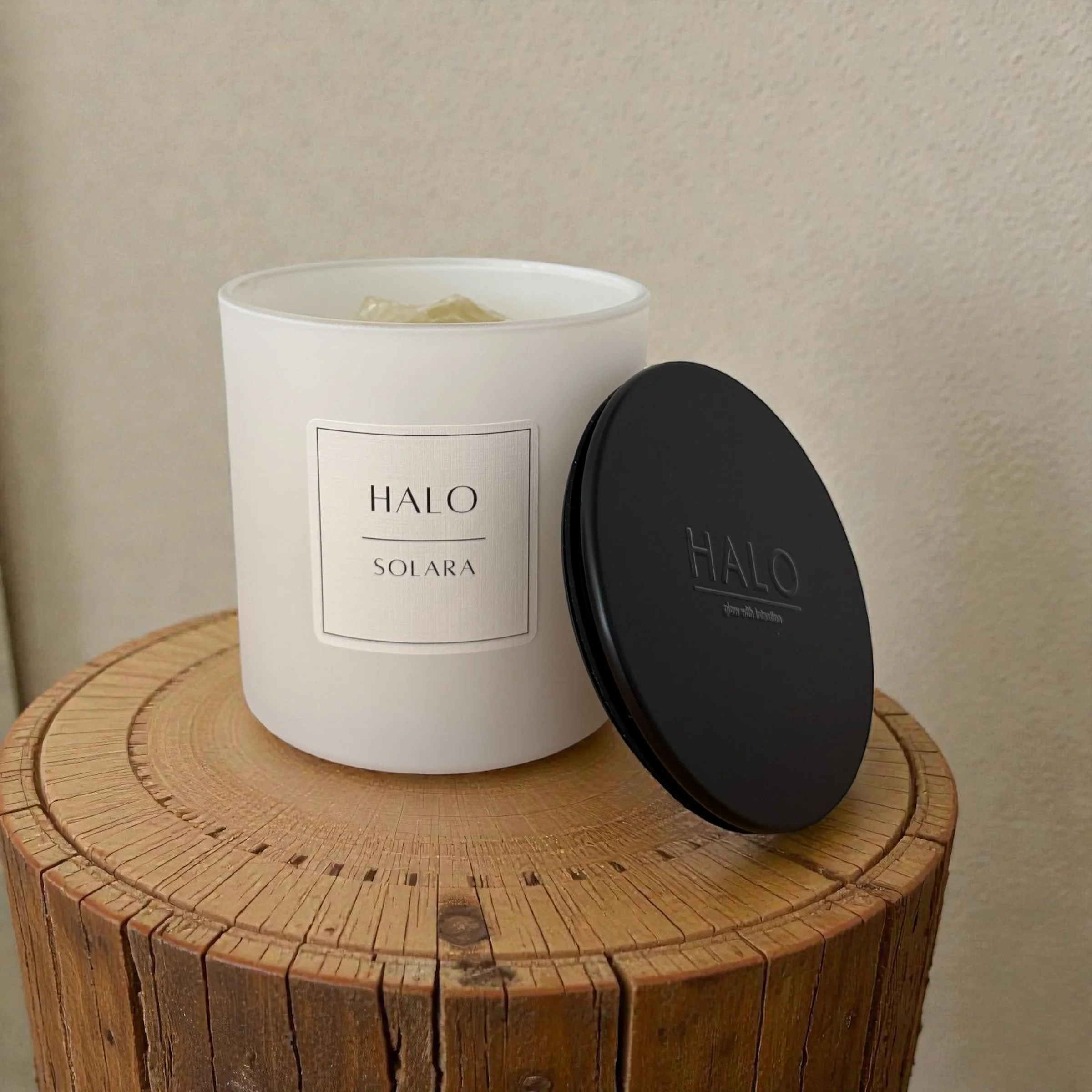 A white matte candle jar labeled 'HALO SOLARA' and a black metal lid also with HALO branding, placed on a round wooden surface against a beige wall. Citrine crystals appear at the top of the candle.