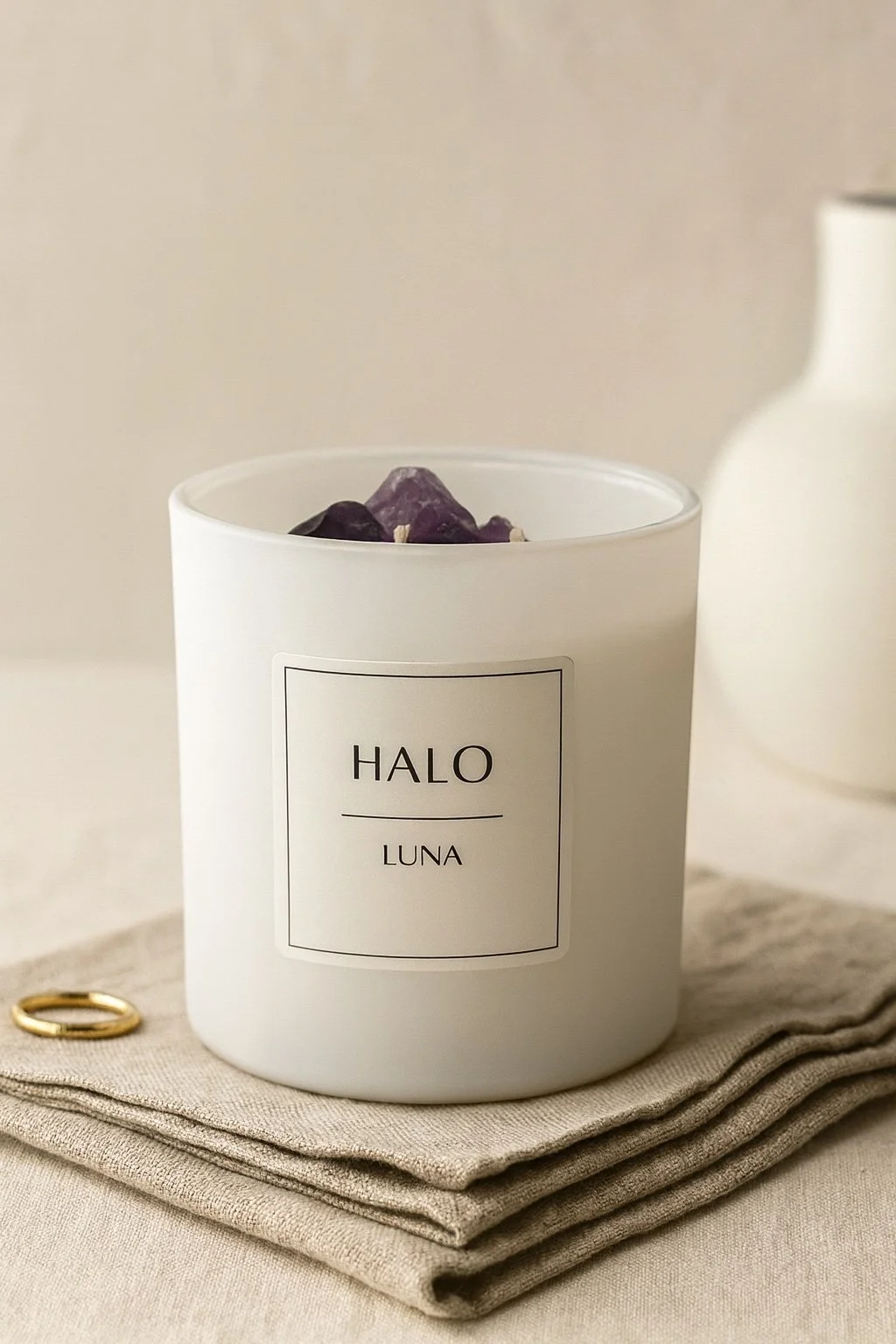 A white candle with a label reading 'HALO LUNA' sits on a folded beige fabric, with purple amethyst crystals inside the candle. A gold ring and a white vase are in the background.