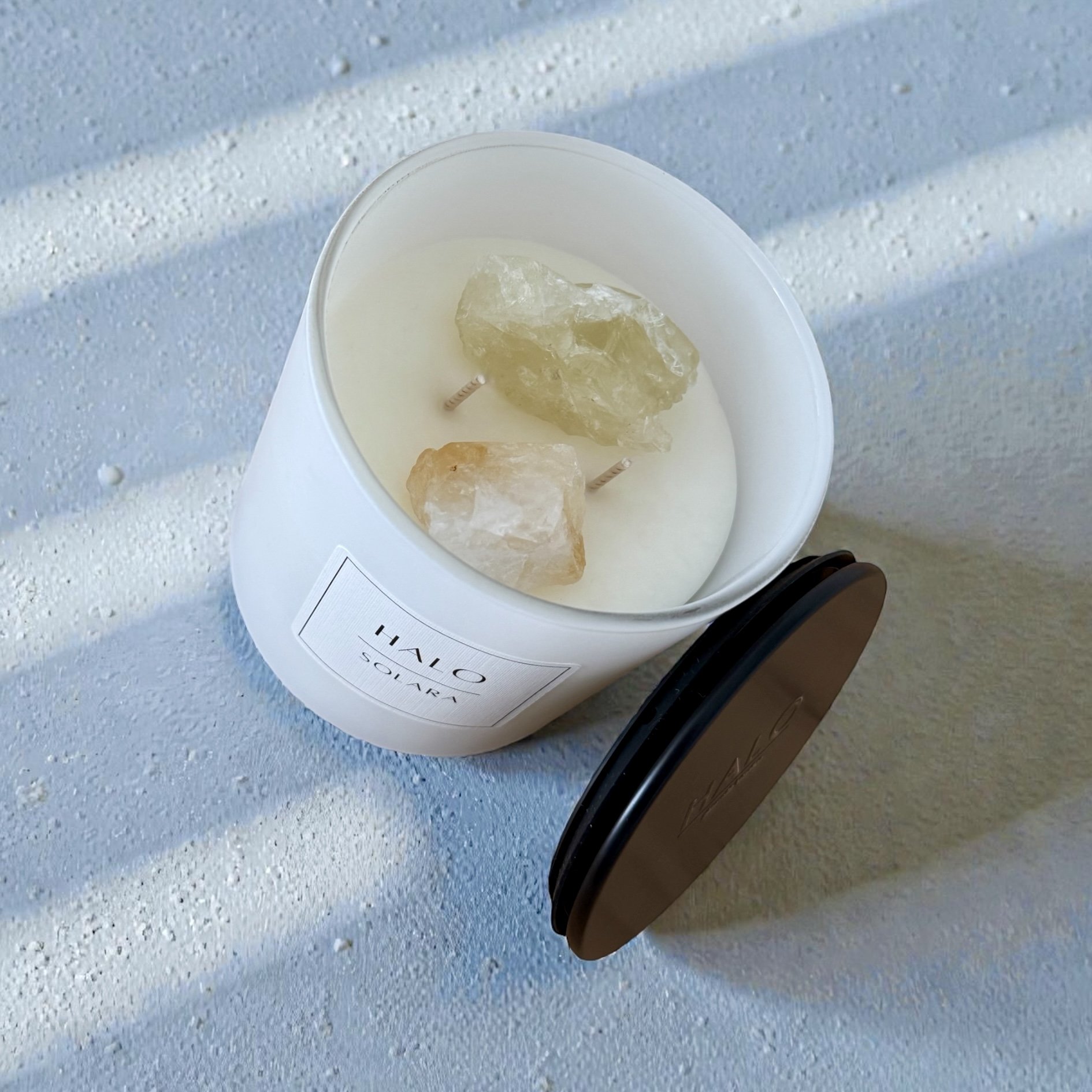 White scented candle with two large, rough citrine crystals on top, placed on a blue textured surface with sunlight streaks, black lid placed beside it, and a label that reads 'HAVO SOLARA' on the candle.
