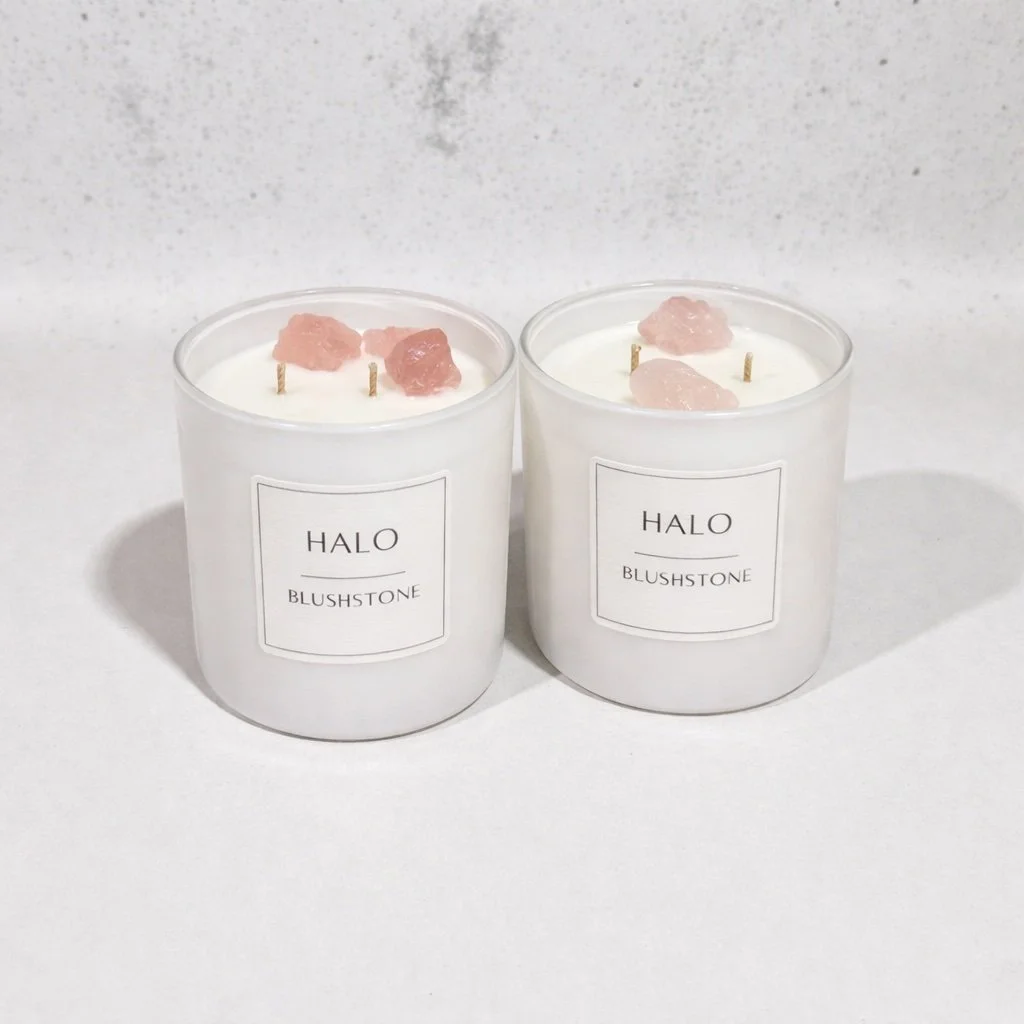 Two white candles labeled HALO BLUSHSTONE with pink rose quartz crystals and cotton wicks inside, placed on a white surface against a white background.