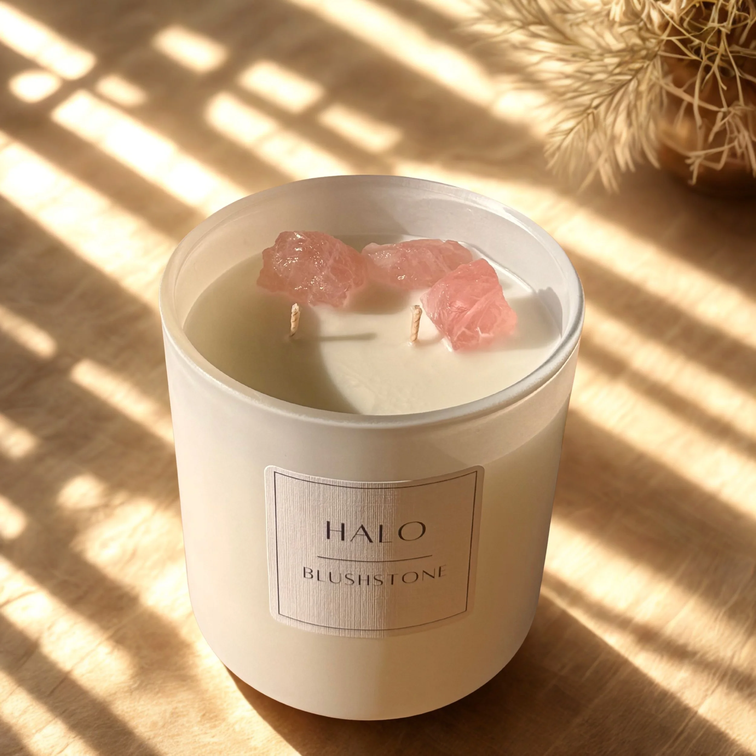 A white candle in a glass jar labeled 'HALO BLUSH TONE' with three rose quartz crystals inside, placed on a beige surface with shadows from blinds and some dried plants in the background.