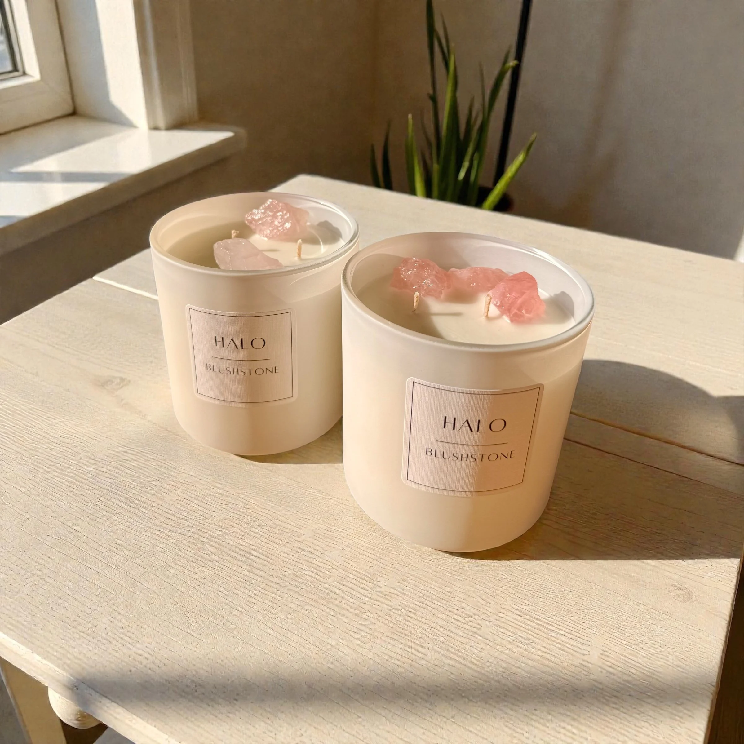 Two white candles in glass containers labeled "HALO BLUSHSTONE" with rose quartz crystals on top, placed on a light-colored wooden surface near a window with sunlight and a green plant in the background.