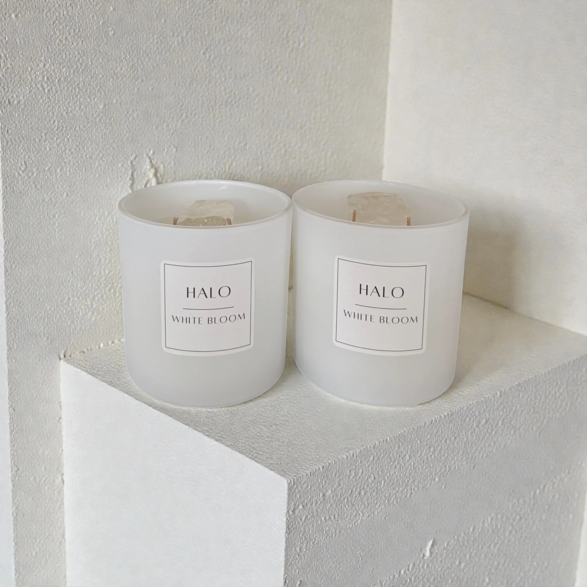 Two white candles in matte frosted glass holders labeled 'HALO White Bloom. Inside there is large clear quartz crystals.