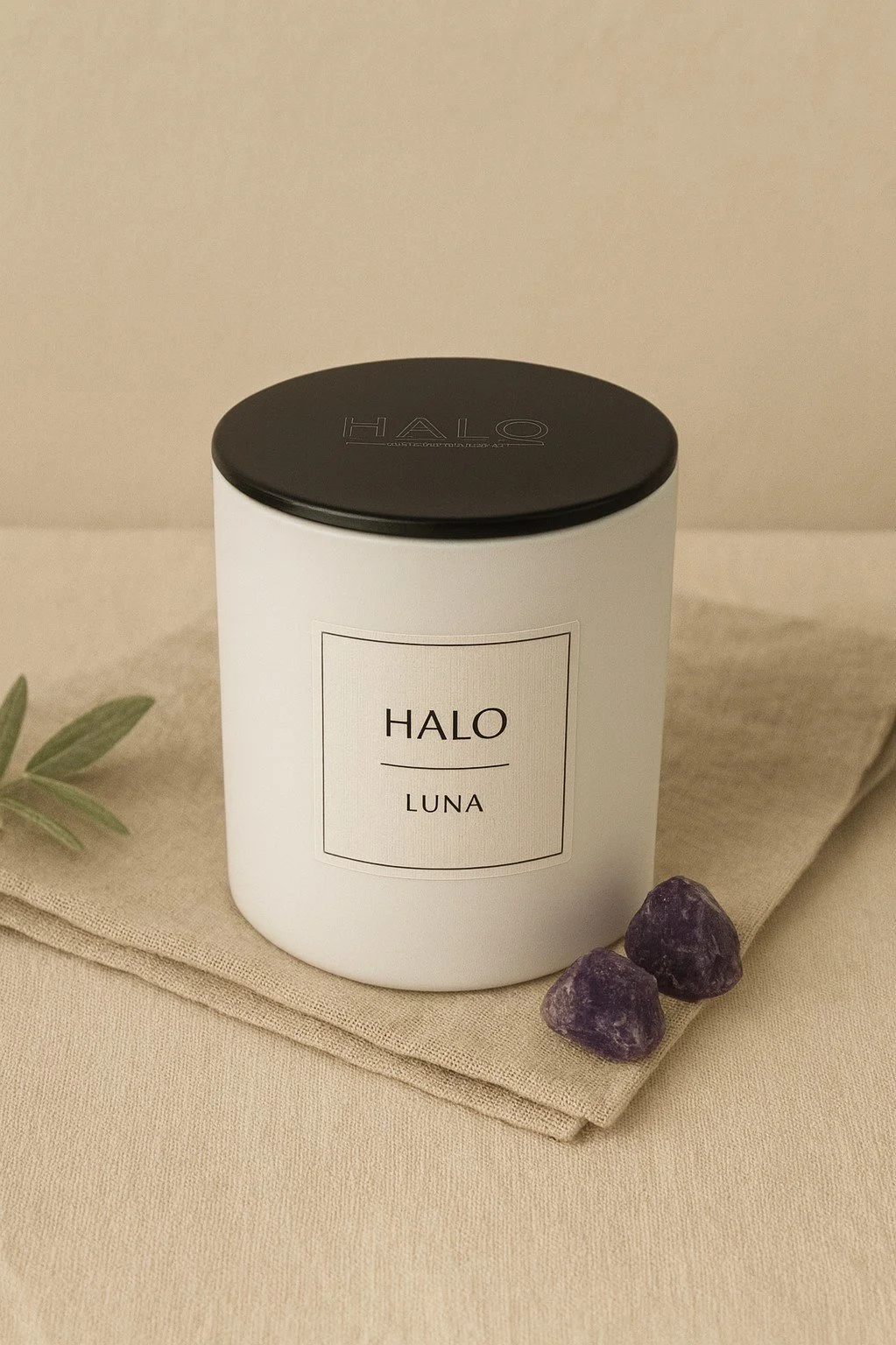 A white candle jar with a black lid labeled "HALO LUNA" placed on a beige fabric surface. There are two purple amethyst crystals, a small branch with green leaves, and a folded beige cloth in the background.
