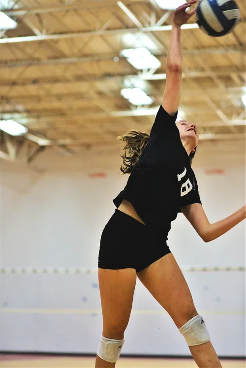 A female volleyball player in a black uniform jumping to hit the ball in an indoor gym.