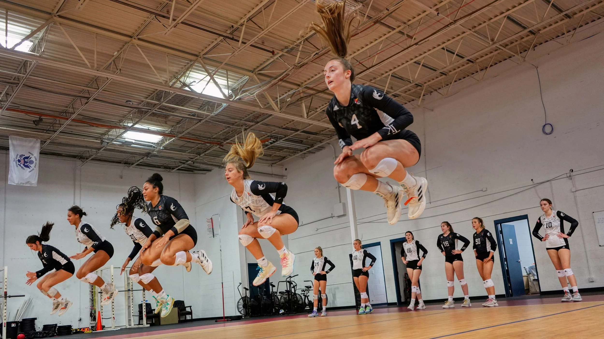 Volleyball team practicing jumps in an indoor gym, with some players airborne and some waiting on the ground observing.