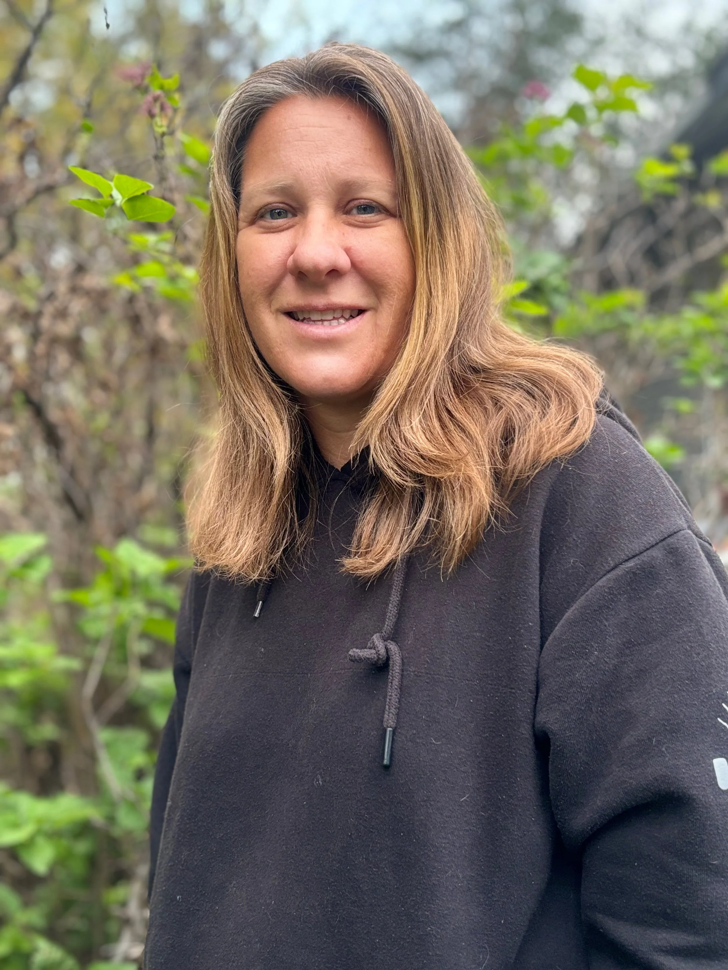 A woman with long light brown hair wearing a black hoodie, smiling outdoors with green foliage in the background.