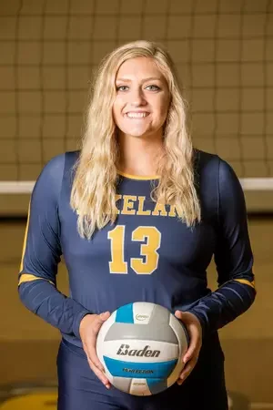 A young woman with long blonde hair, wearing a navy blue volleyball uniform with the number 13, holding a Baden volleyball, standing in an indoor volleyball court.