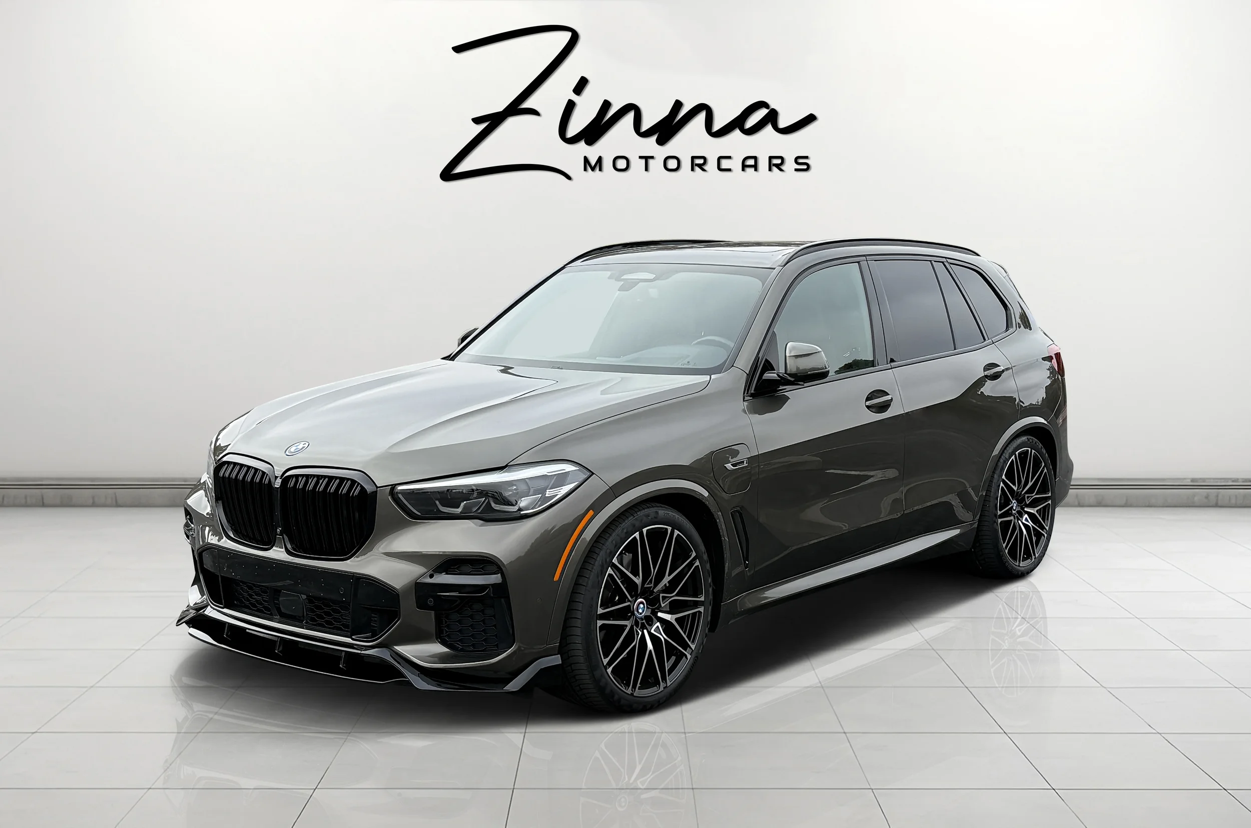 A black and gray BMW SUV parked indoors against a white wall with a sign that reads "Zinna Motorcars."