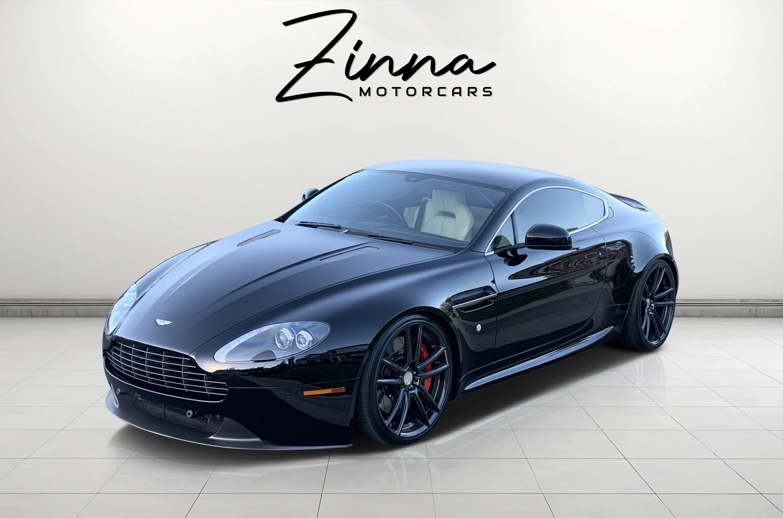 Black Aston Martin sports car on showroom floor with Zinna Motorcars logo on white wall behind it.