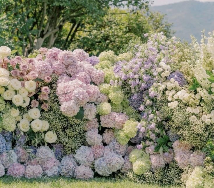 Colorful floral arrangement of pink, purple, white, and green hydrangeas and roses outdoors with trees and mountains in the background.