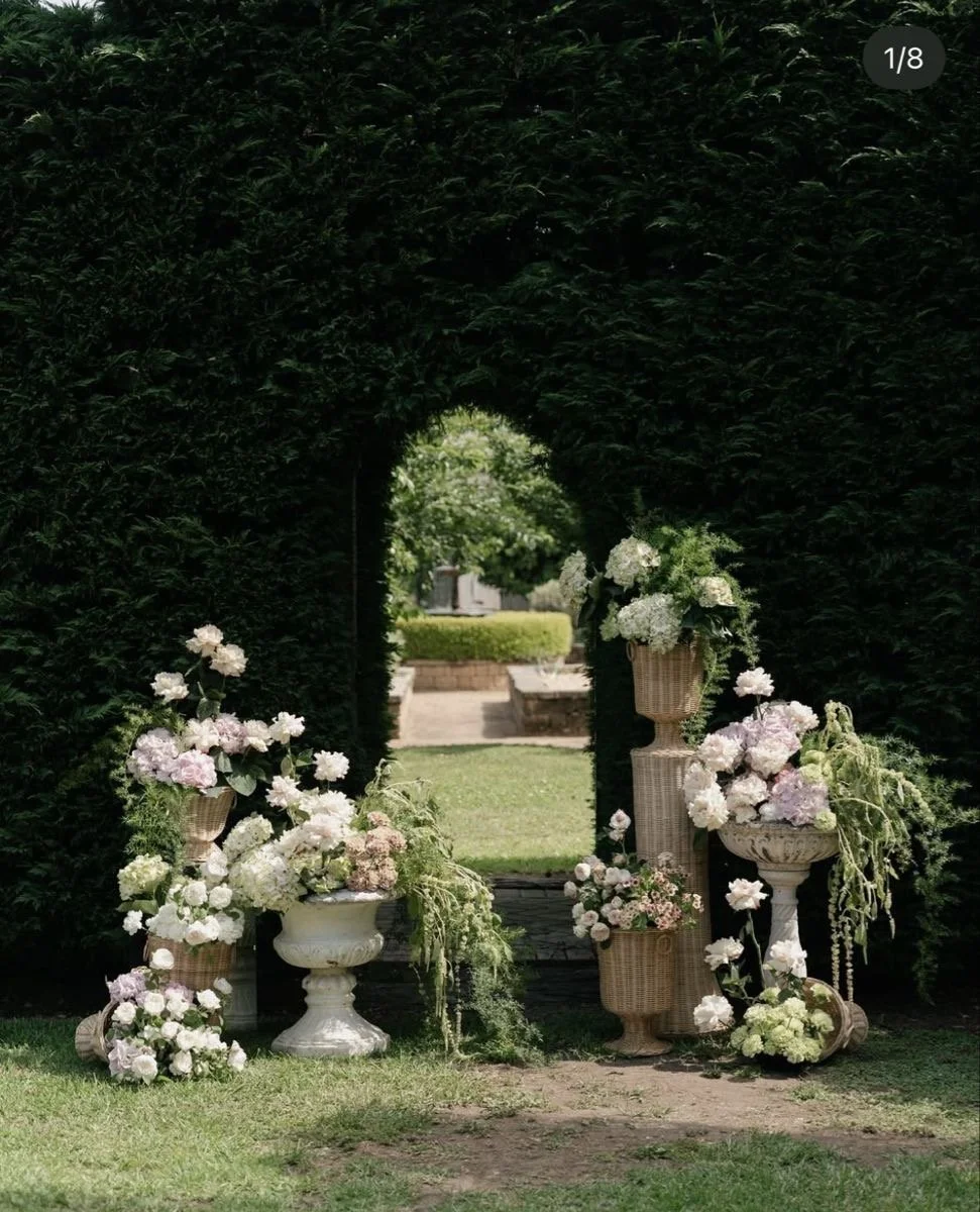 A floral arrangement with white and pale pink flowers in large urns and baskets, arranged outdoors under a hedge archway with a garden in the background.