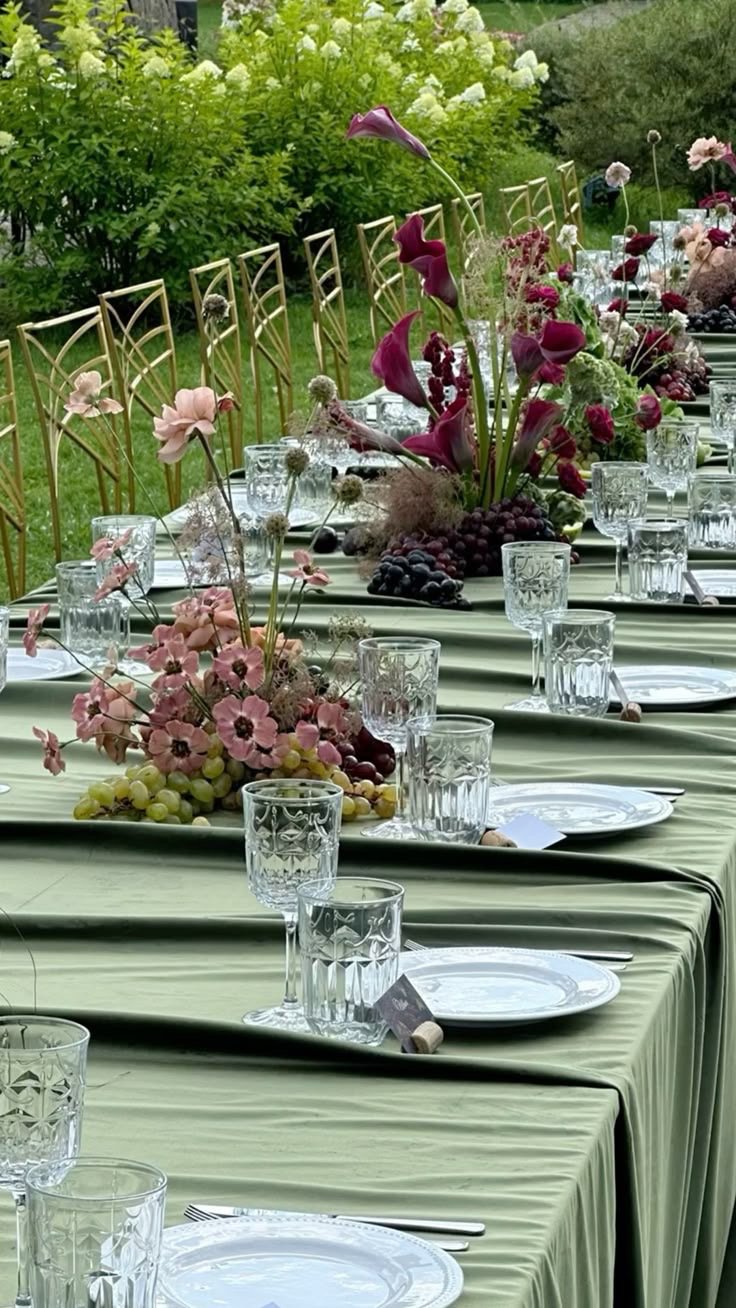 An outdoor dining table set with plates, glasses, and floral centerpieces on a green tablecloth, surrounded by gold chairs, with greenery and trees in the background.