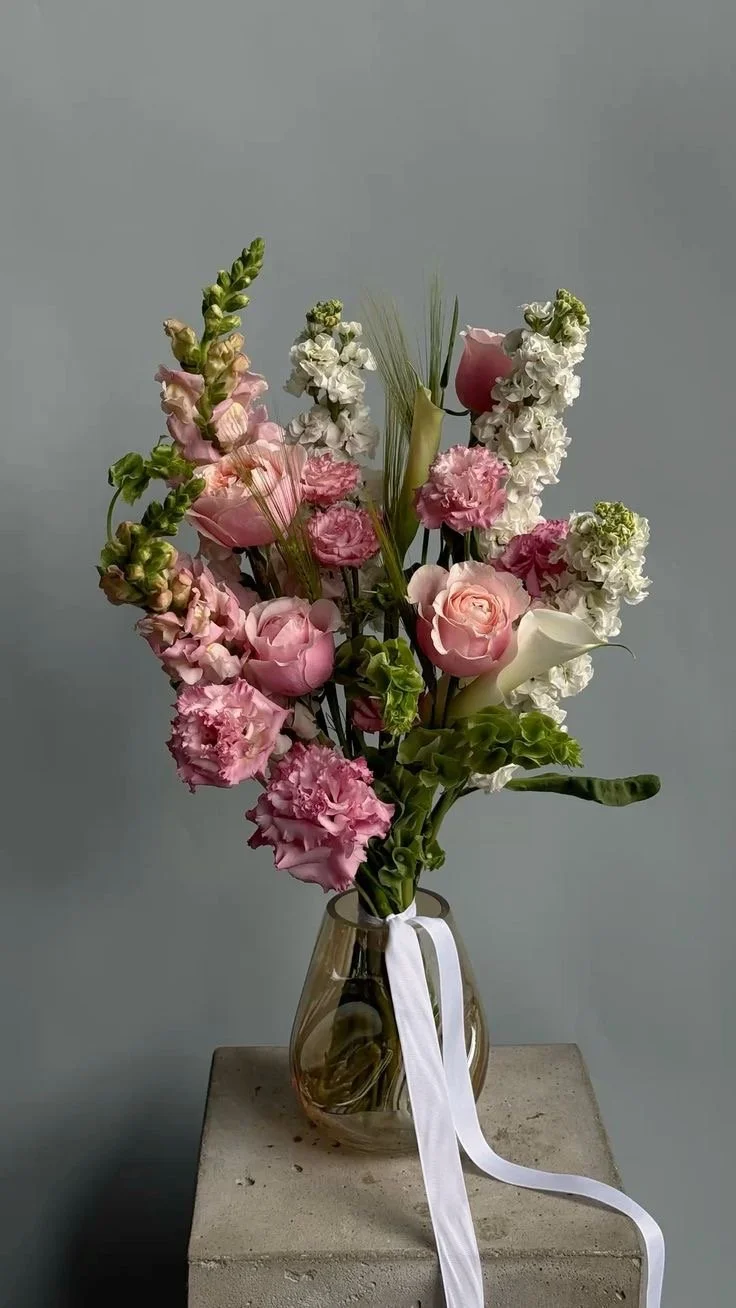 A glass vase with a white ribbon tied around the neck, holding a bouquet of pink, white, and green flowers on a concrete surface against a plain gray background.