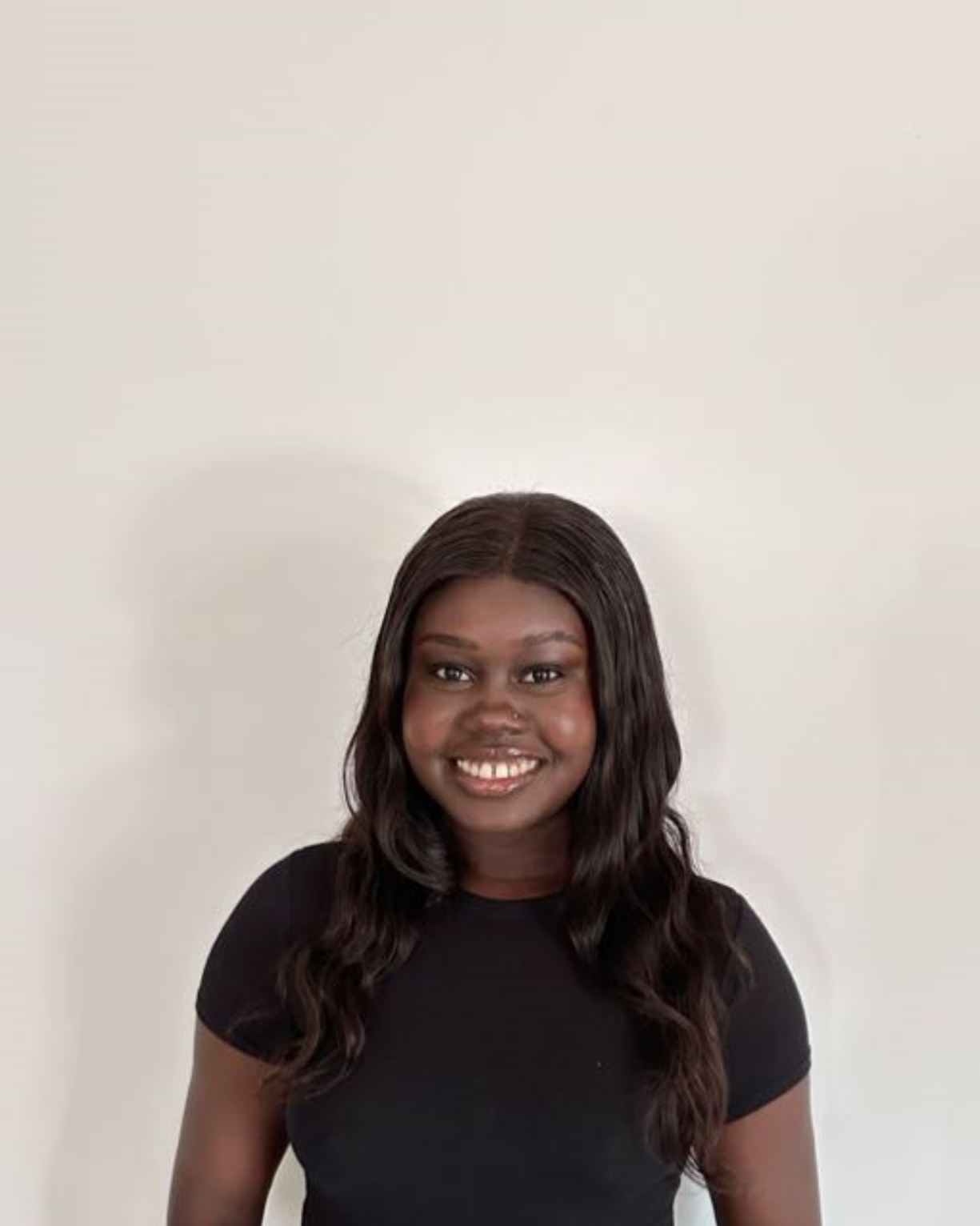 A young woman with dark skin, long curly hair, wearing a black t-shirt, smiling, standing against a plain white wall.