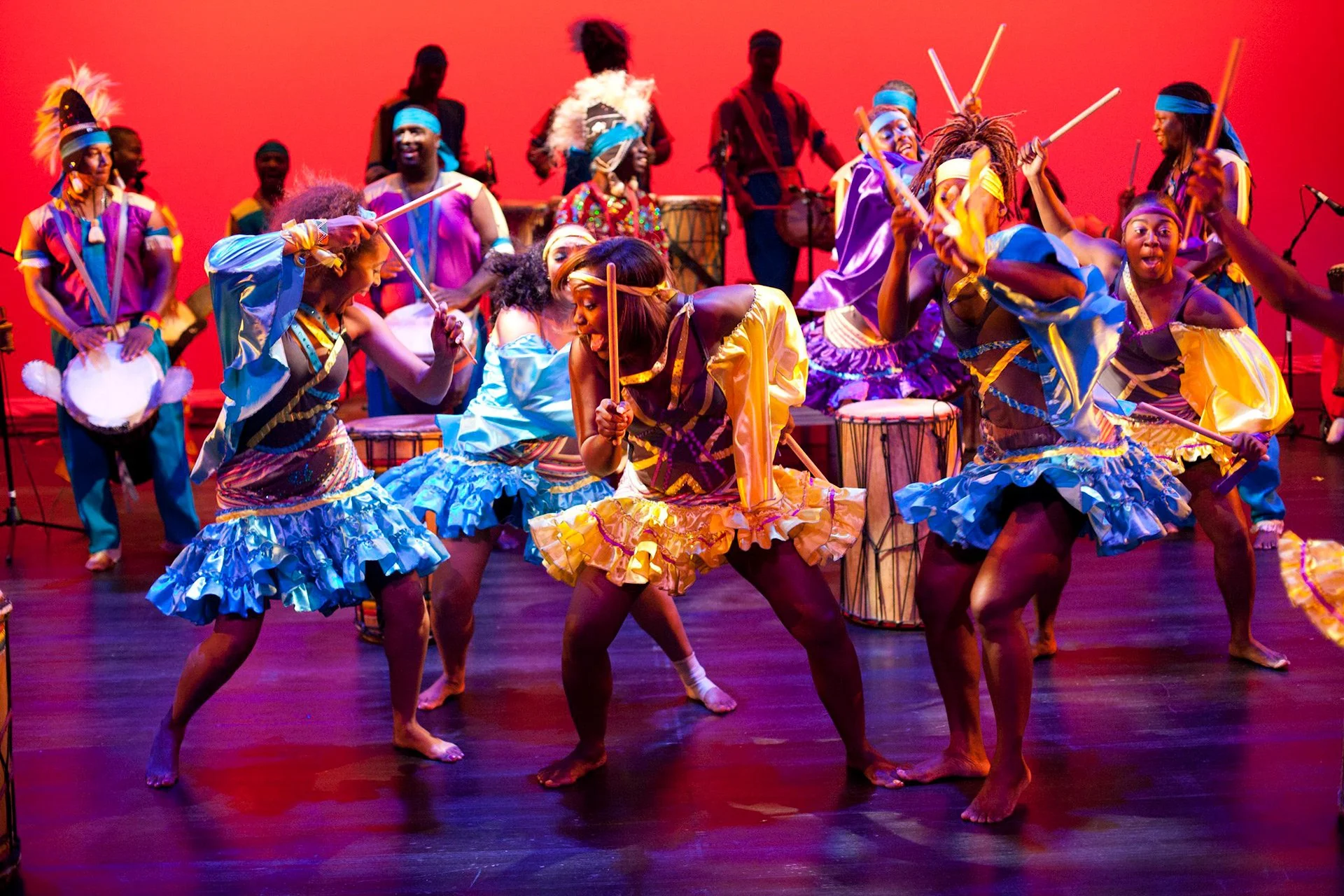 Group of dancers in colorful costumes, performing with drums and sticks on stage with a red background.