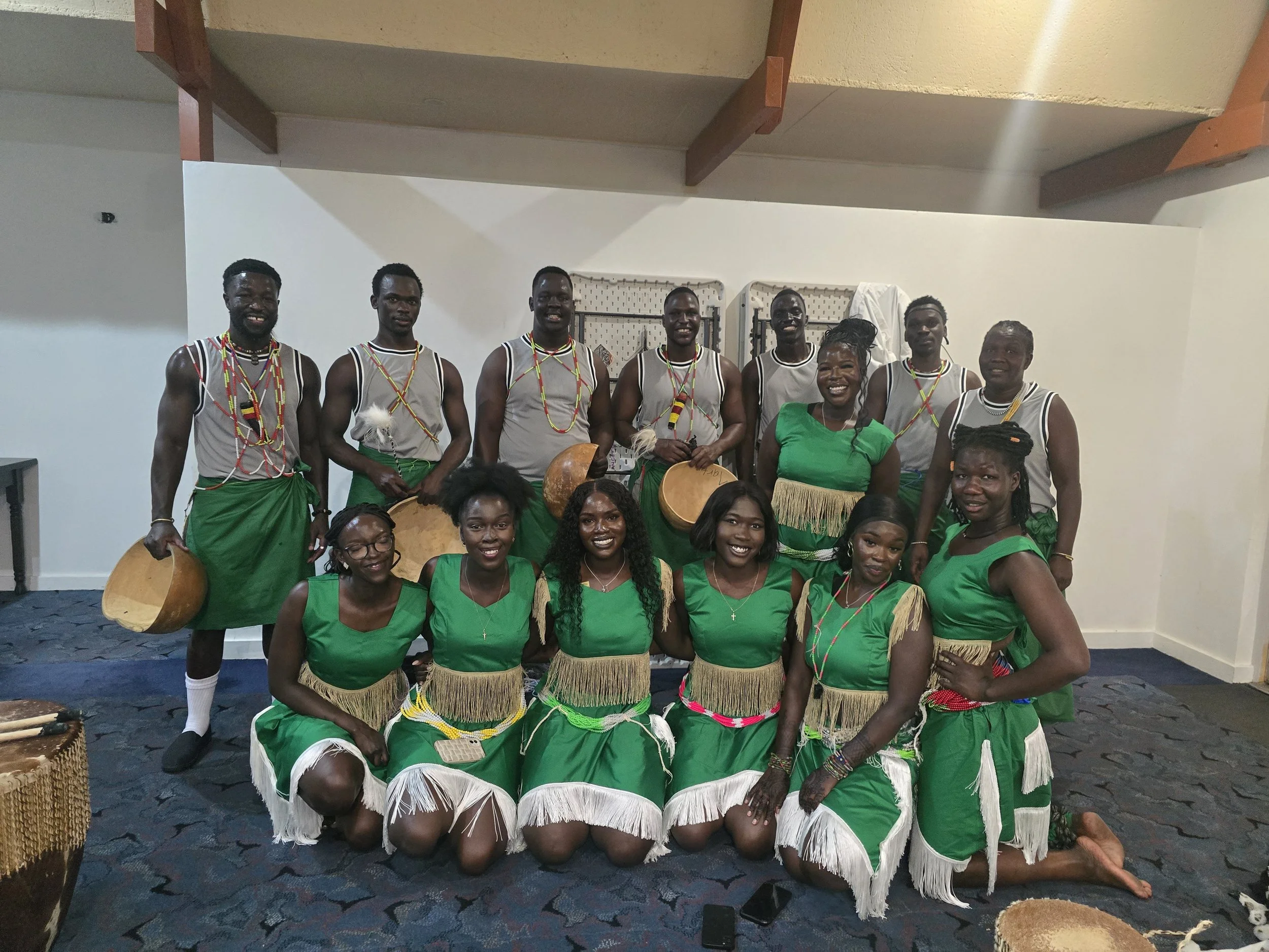A group of men and women in traditional African attire, posing indoors. The men are standing in the back row, wearing gray tops, green skirts, and beaded necklaces. Some are holding drums. The women are kneeling in front, dressed in green dresses with fringe and white details, smiling at the camera.