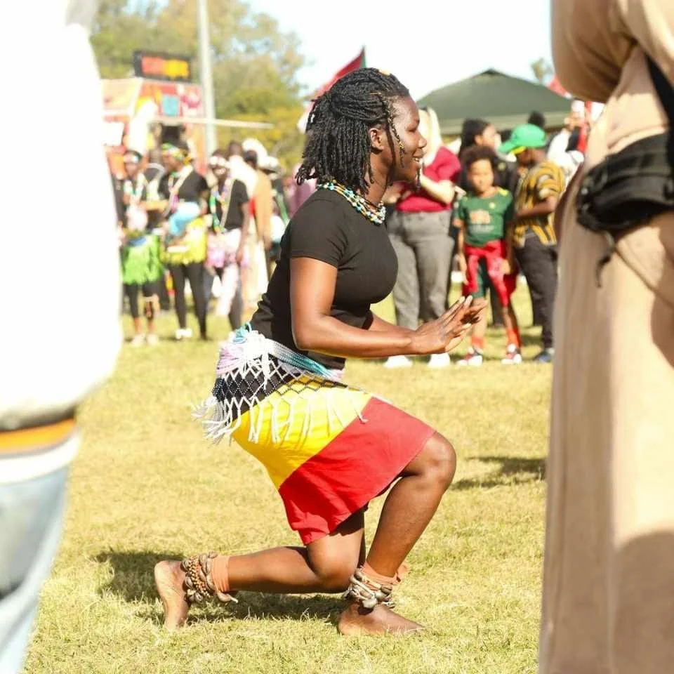 A woman kneeling on the grass at an outdoor festival or gathering, surrounded by other people. She is wearing traditional clothing and jewelry, including ankle beads, and is smiling.