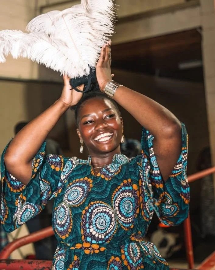A woman wearing a colorful, patterned dress is smiling and holding a large feathered headdress or fans above her head.