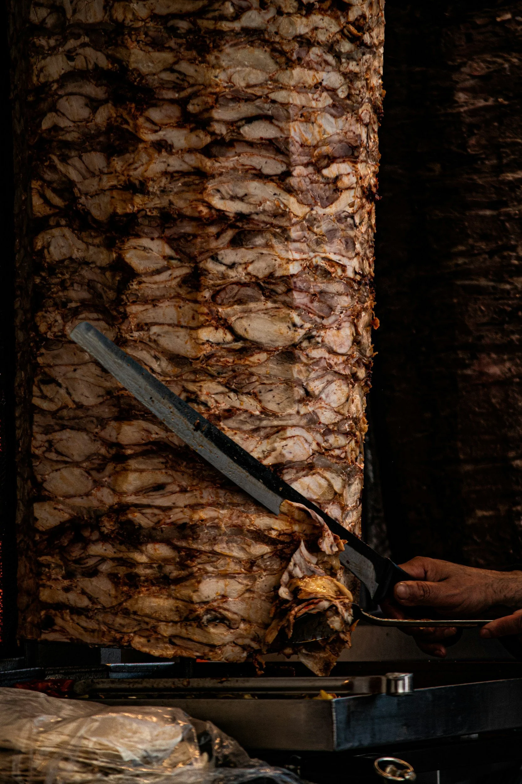 Close-up of a vertical spit roast with sliced meat, used for making gyros or shawarma, with a person carving the meat using a large knife.