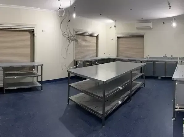 Empty industrial kitchen with stainless steel work tables, closed window shutters, and hanging wires on the wall.