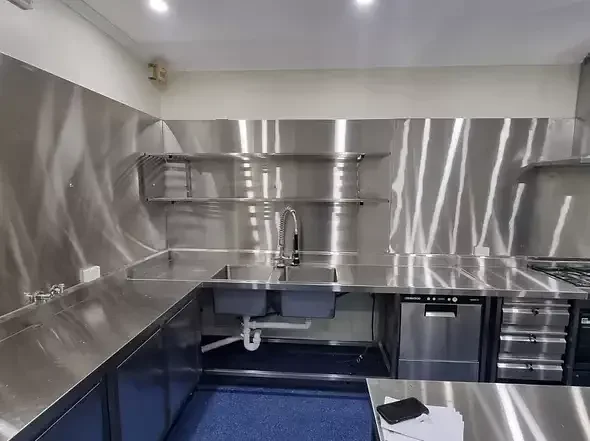 Stainless steel commercial kitchen with sink, open shelving, and a stove.
